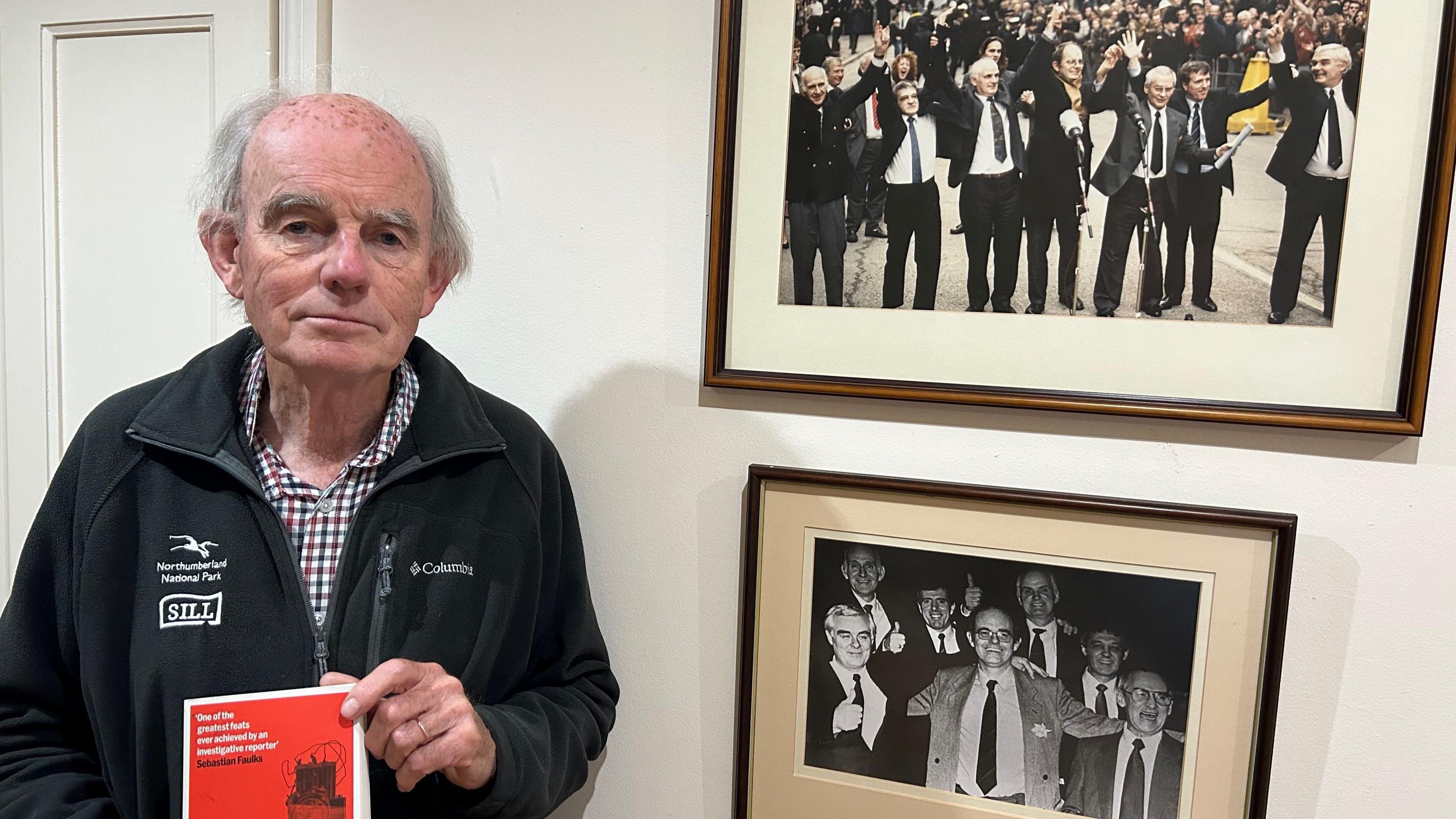 Chris Mullin stands facing the camera. He has grey hair and is wearing a checked shirt and black fleece, and holding a book with a red cover. To his left are framed black and white photographs of himself and the Birmingham Six, celebrating their release.