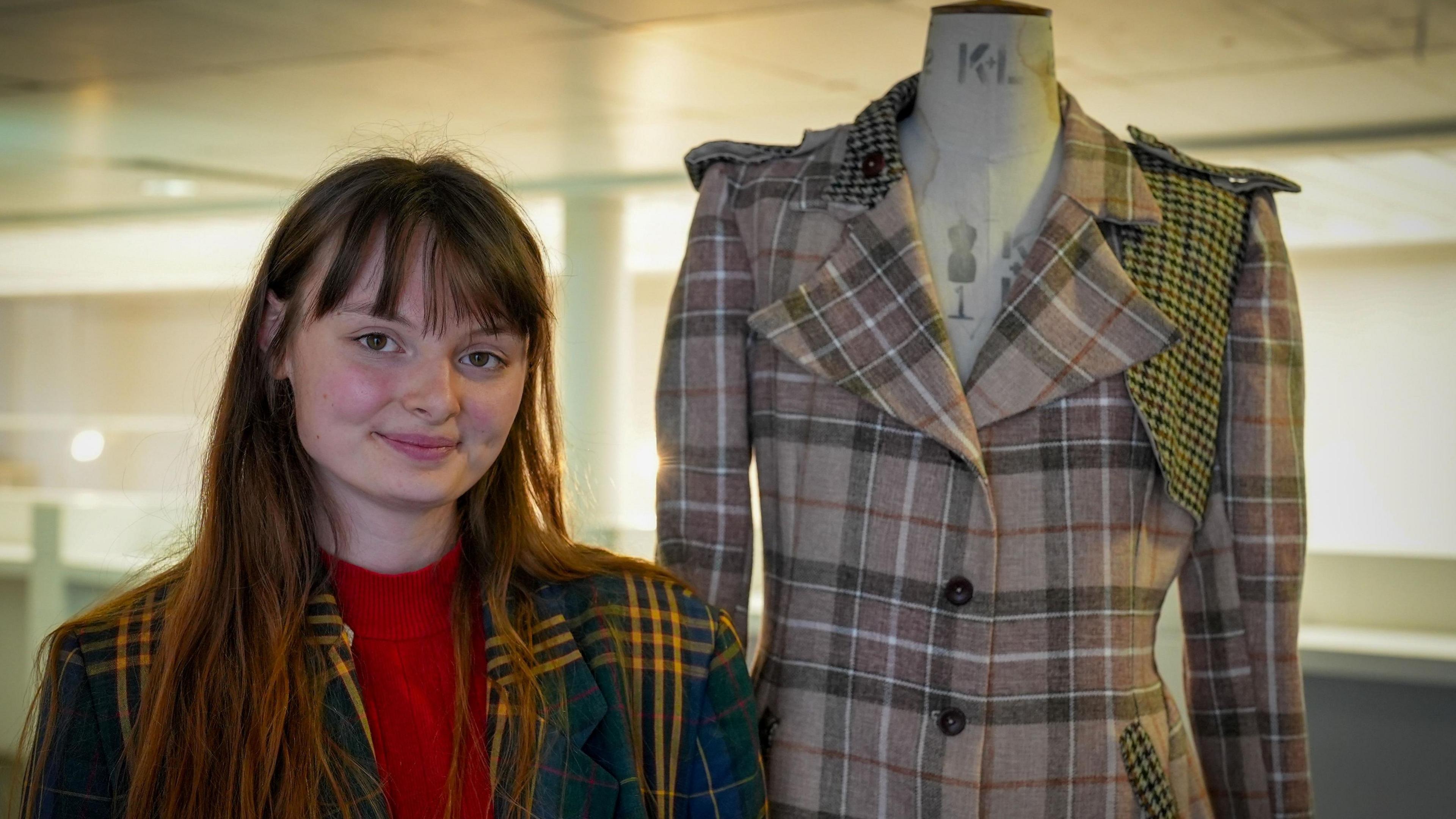 Milly Toombes, a woman with a fringe and wearing a green, blue and yellow tartan coat, smiles. Next to her is a mannequin wearing a beige tartan coat