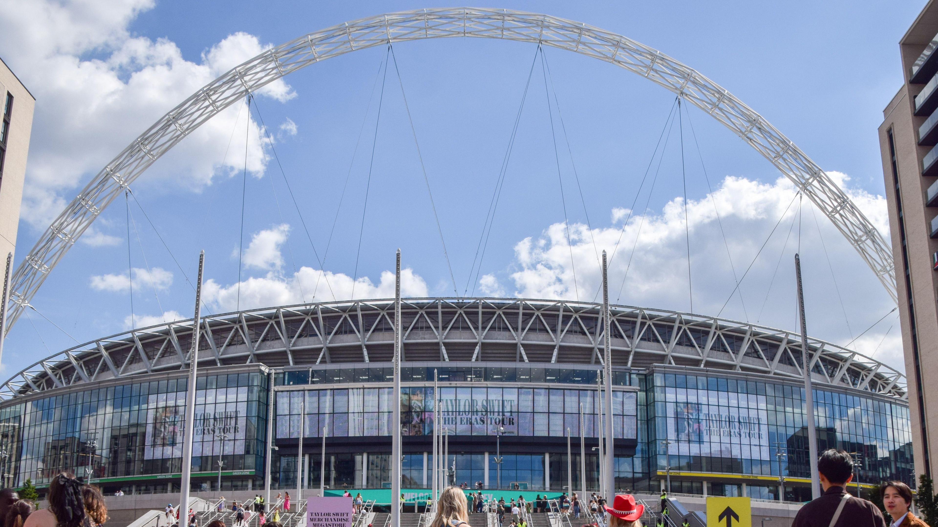 People gather outside Wembley Stadium, with its arch, on a sunny day.
