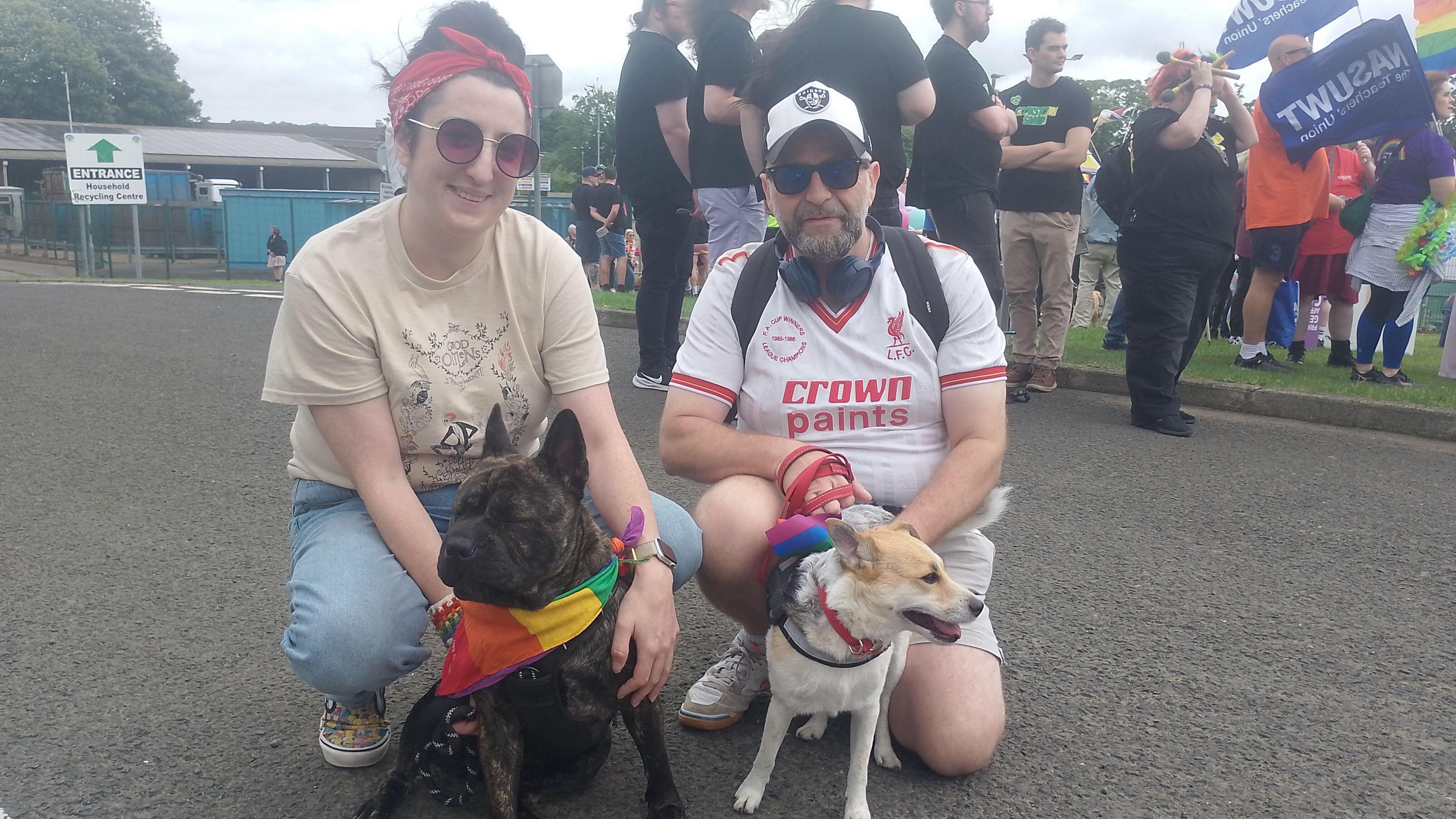 Danni and Brian Penny crouched down on the ground with their two dogs. Danni has dark hair tied up with a red bandana, wearing sunglasses, a beige t-shirt and jeans. Brian is wearing a white cap, sunglasses, headphones around his neck, a Liverpool jersey and shorts. One of the dogs is a dark brown staffy wearing a rainbow bandana, the other dog is white and light brown with a red collar.