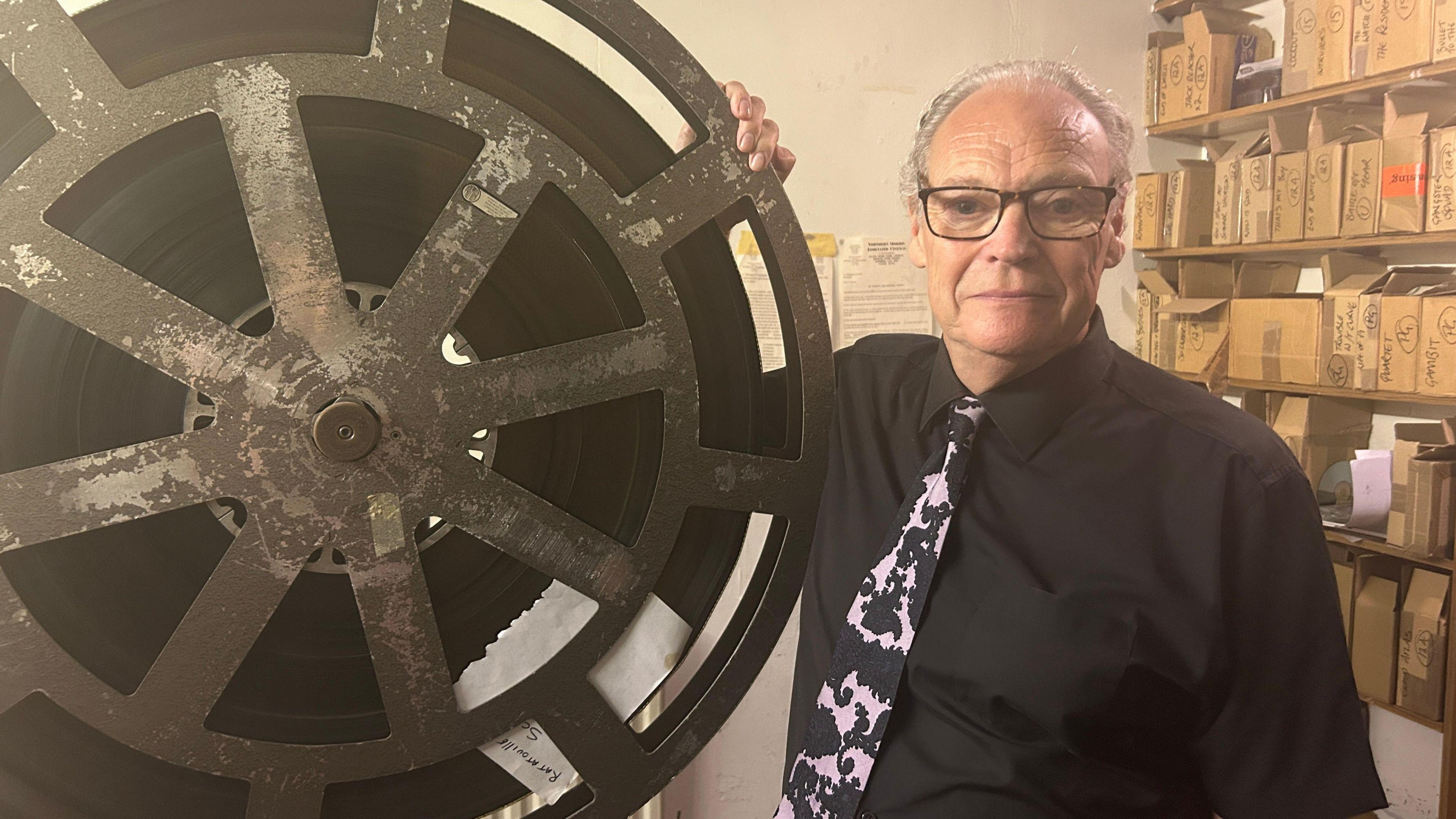 An older man standing next to a giant projectionist's wheel of film. He is surrounded by boxes on shelves.