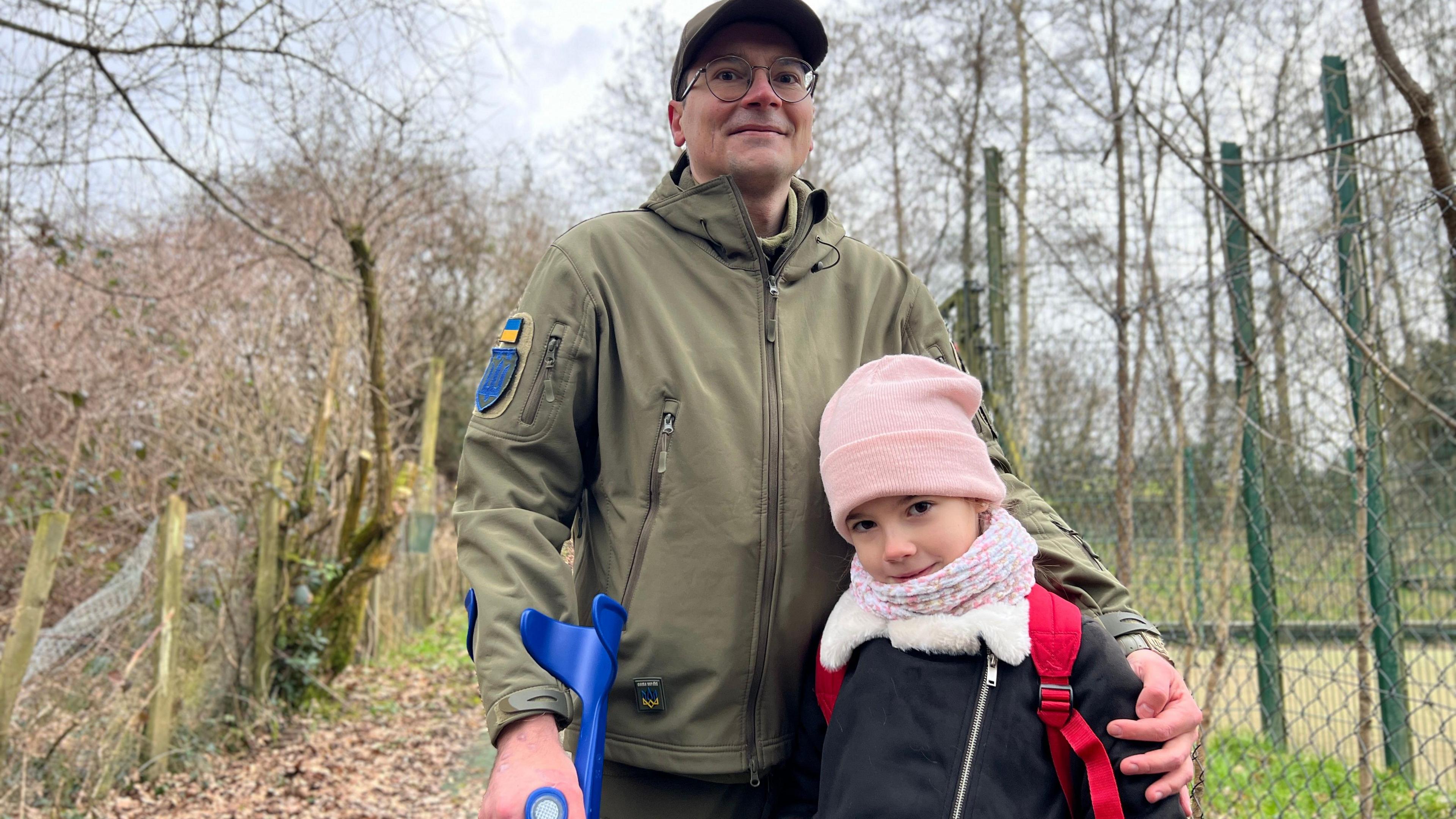 A man with his left arm around a young girl is looking at the camera. The man is dressed in a khaki green jacket. He is wearing glasses and you can see a small Ukrainian flag on his right arm. The young girl is wearing a pink hat and scarf. You can see the straps of the red school bag she has on her back.  You can see the top of a blue crutch the man is holding in his right hand.