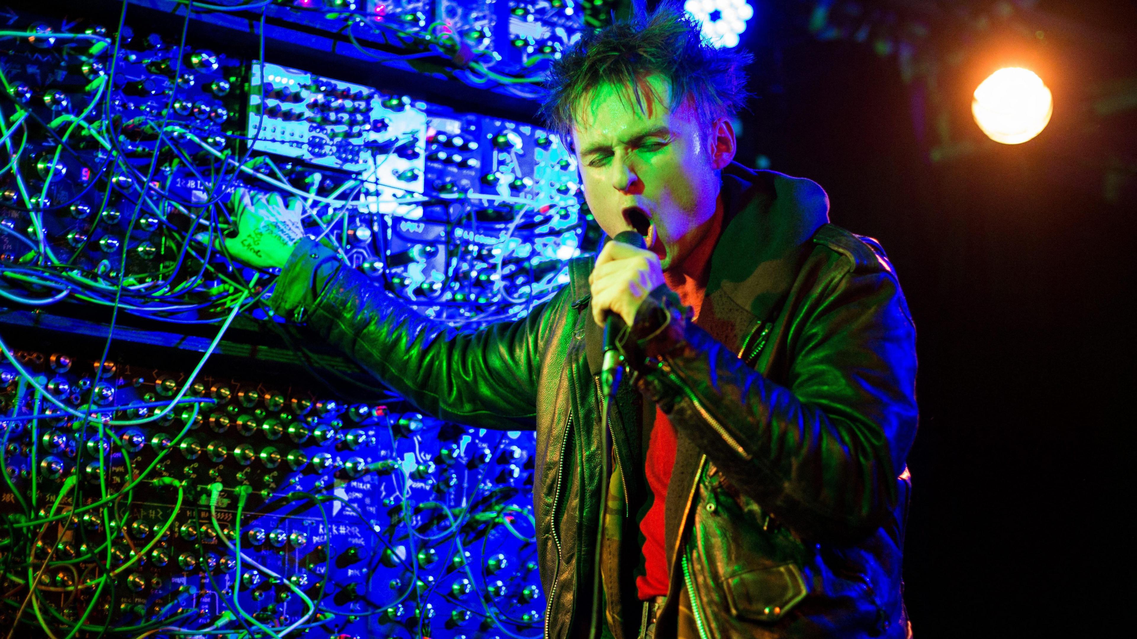 Sam Battle pictured on stage in Oslo, Norway, in 2019, singing into a microphone and with one hand on a very large bank of knobs, dials and wires next to him