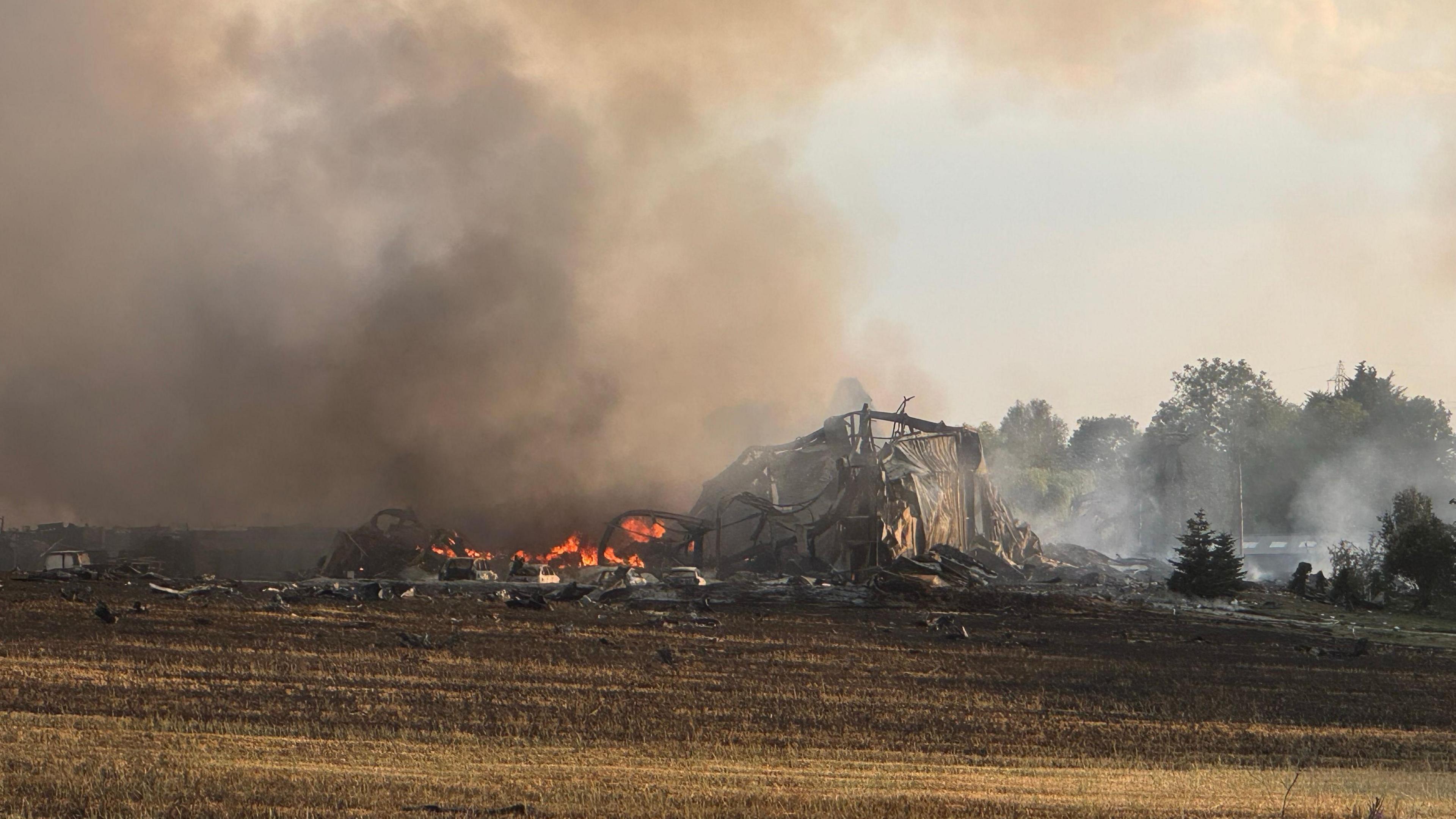 A collapsed building surrounded by fire and smoke on a field.