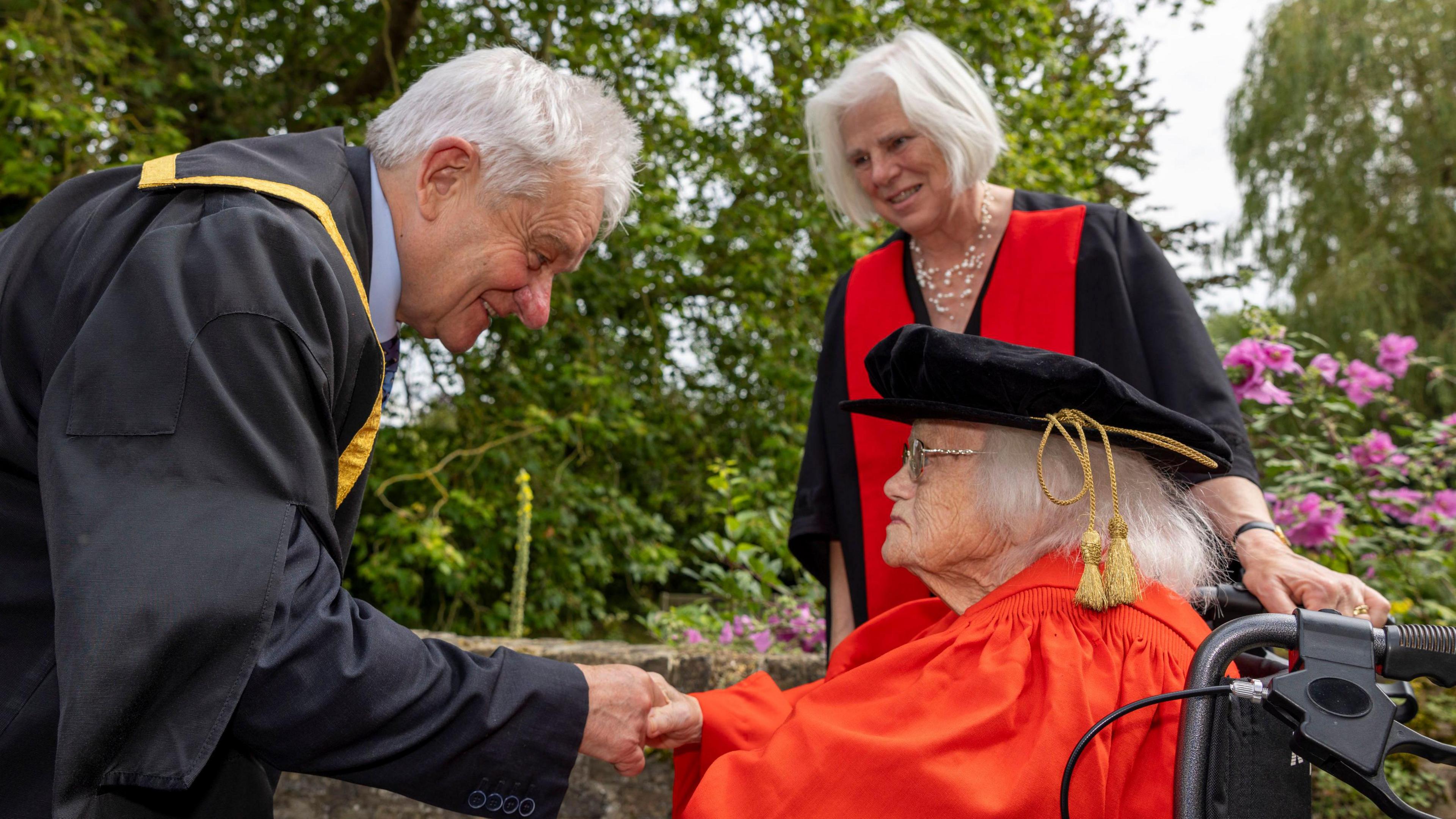 A man dressed in black graduation robes shaking the hand of a woman sitting in a wheelchair wearing red graduation robes