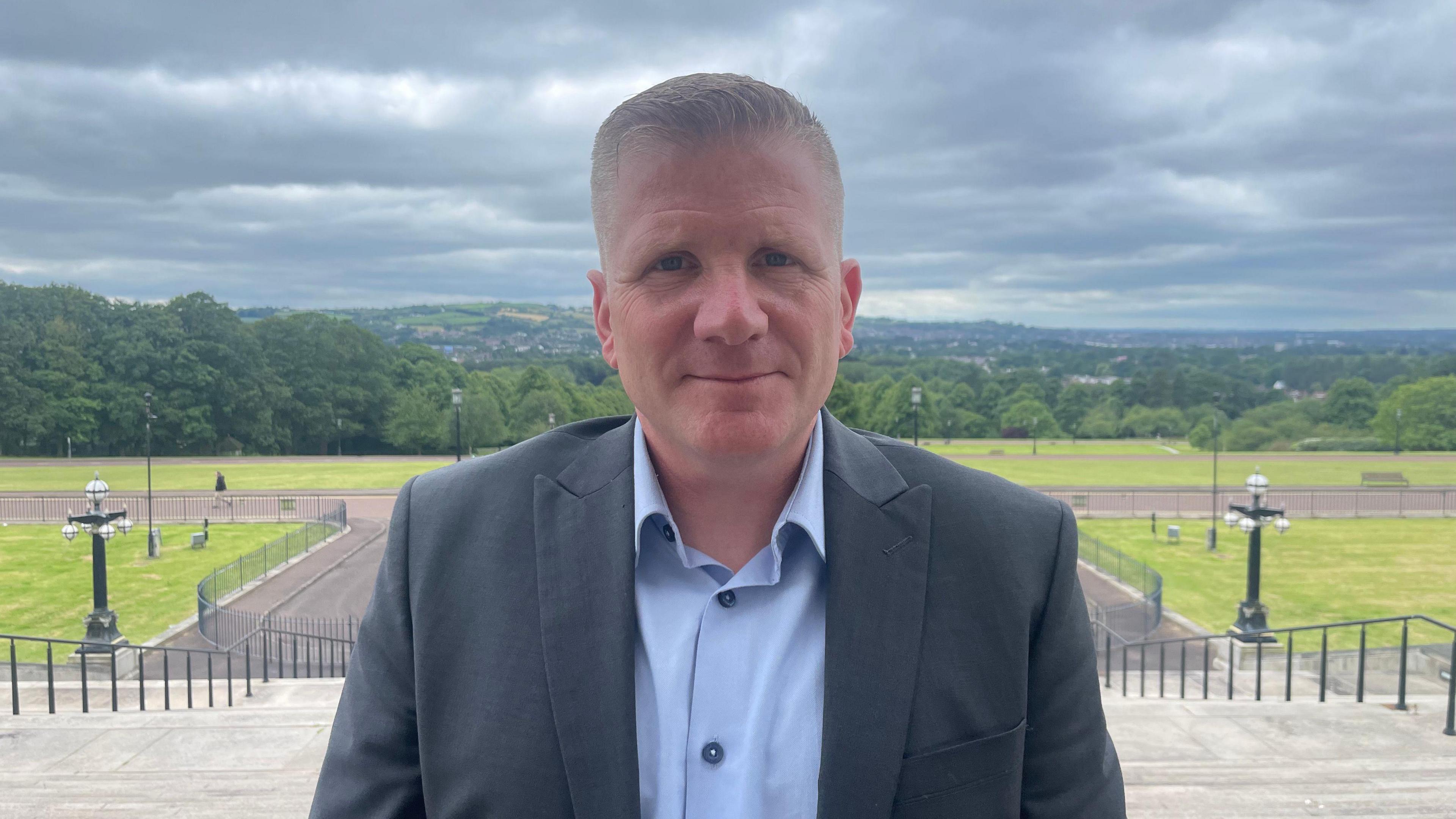 Chris Quinn is standing looking into the camera with a slight smile. He is standing outside with grass and trees in the background. He is wearing a shirt and jacket