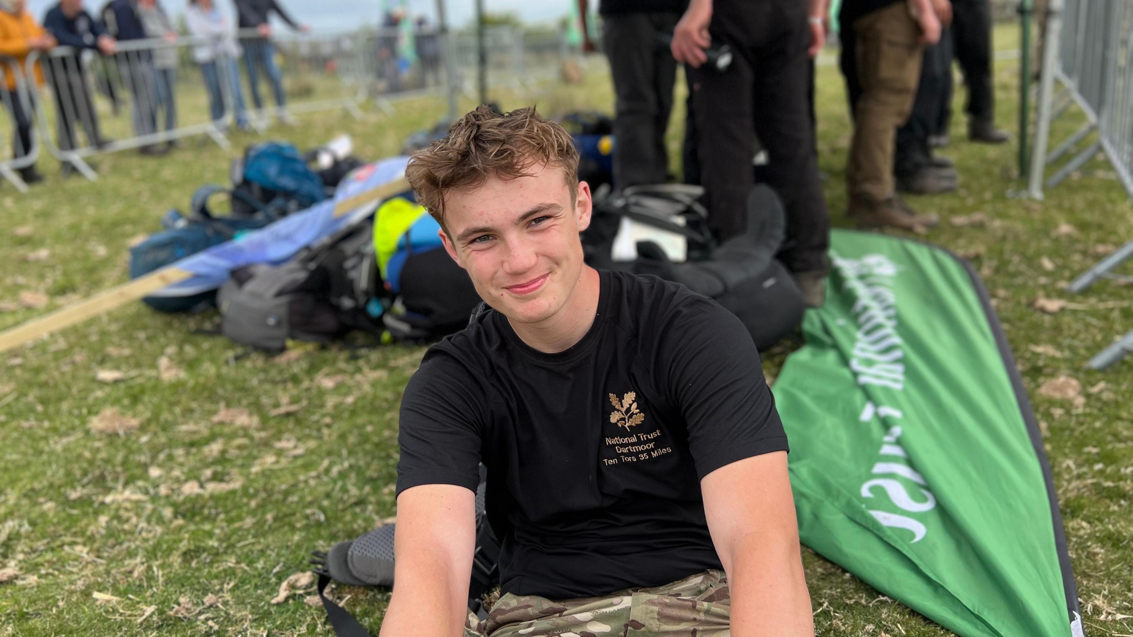 Charlie Snell sat on the floor at the finish line of the Ten Tors Challenge. There are people in the background and rucksacks on the floor next to him.