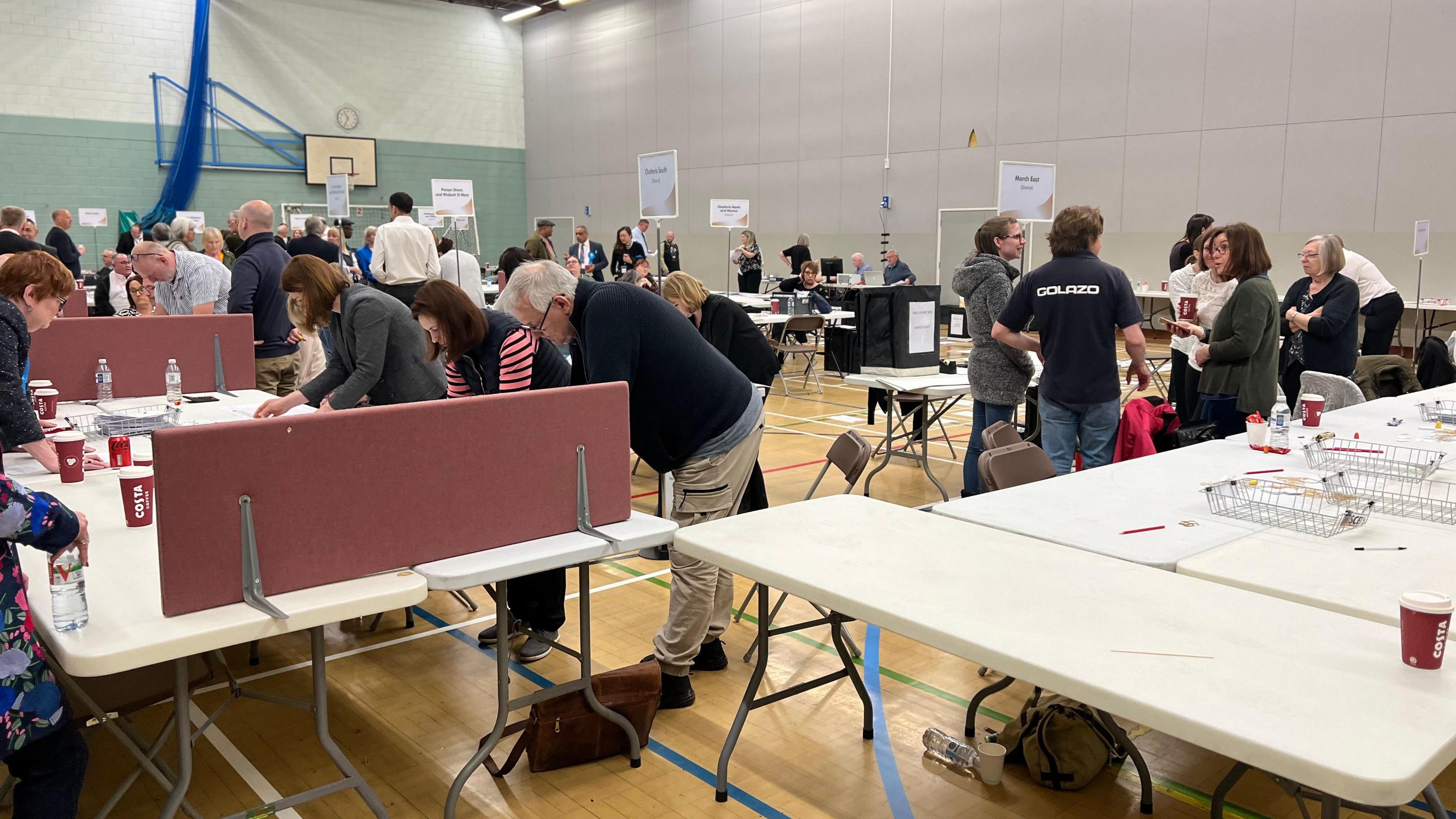 Image shows many people bending over tables as they count ballots in Hudson Leisure Centre's sports hall. 