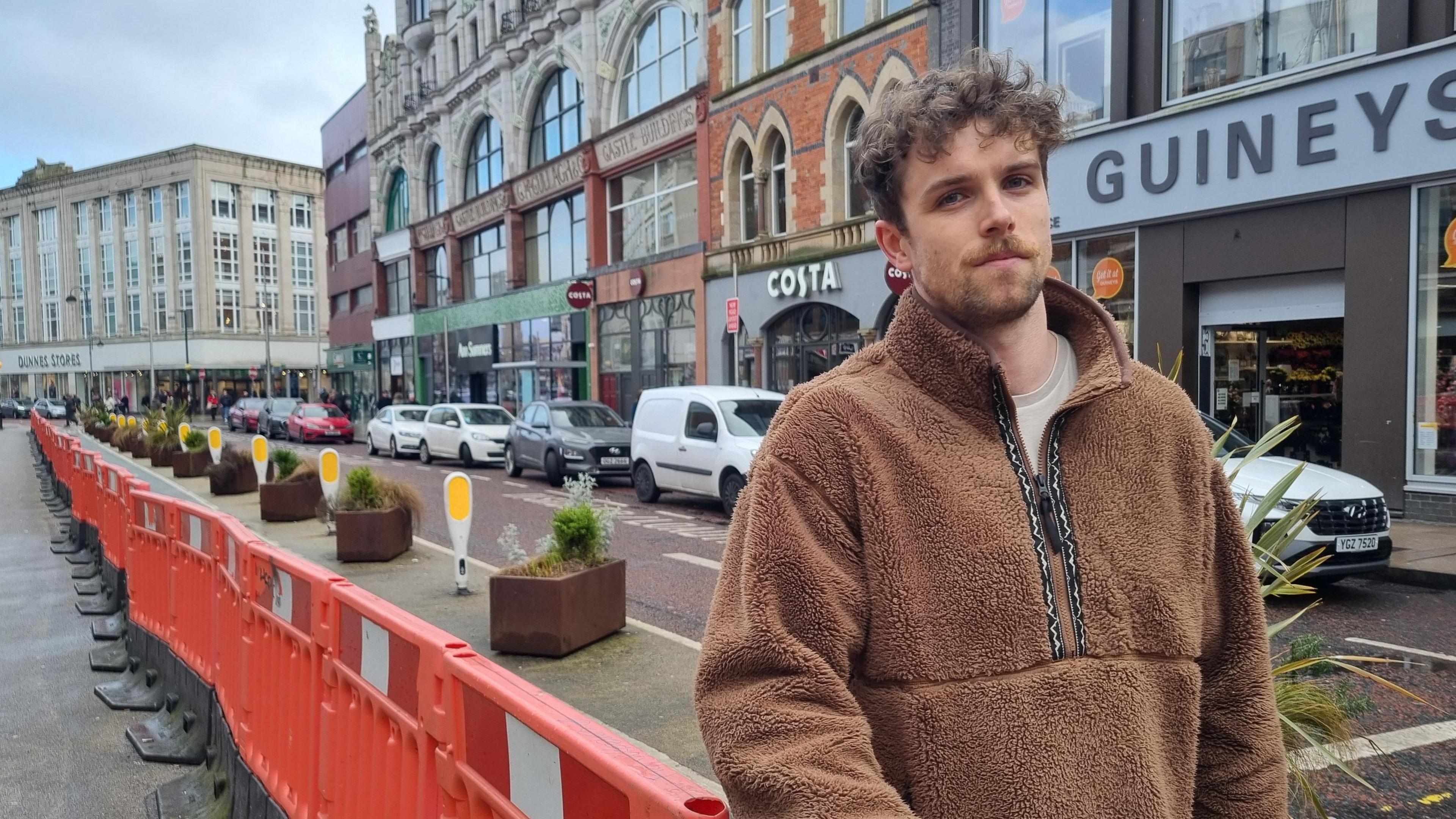 Man wearing a brown teddy material fleece standing in front of an orange temporary barrier along the street. Shops and cars line the other side of the road. 