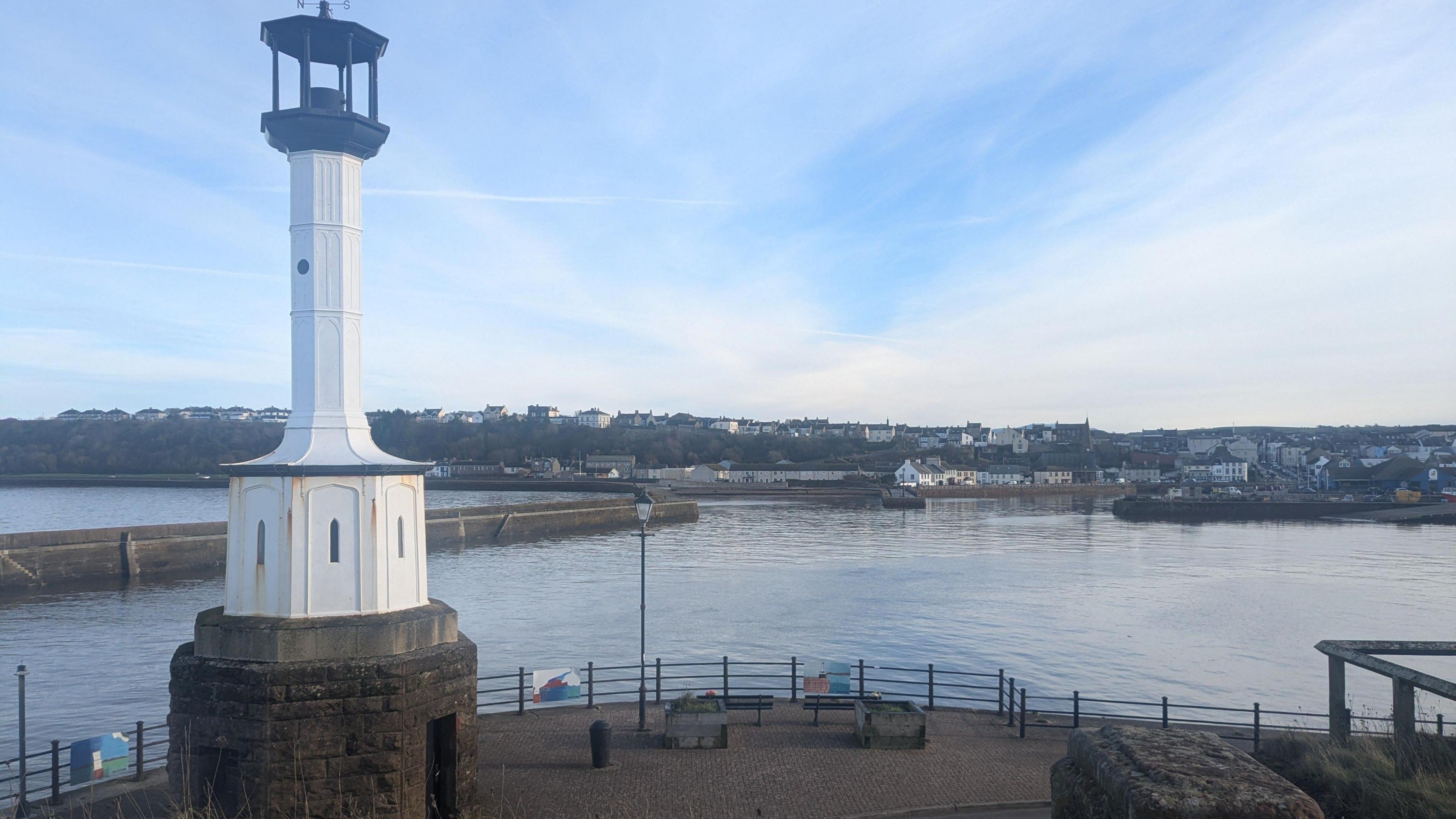 A general view of Maryport, Cumbria, from the lighthouse by the harbour. The white lighthouse is in the foreground, and houses and buildings are visible in the distance over the water.