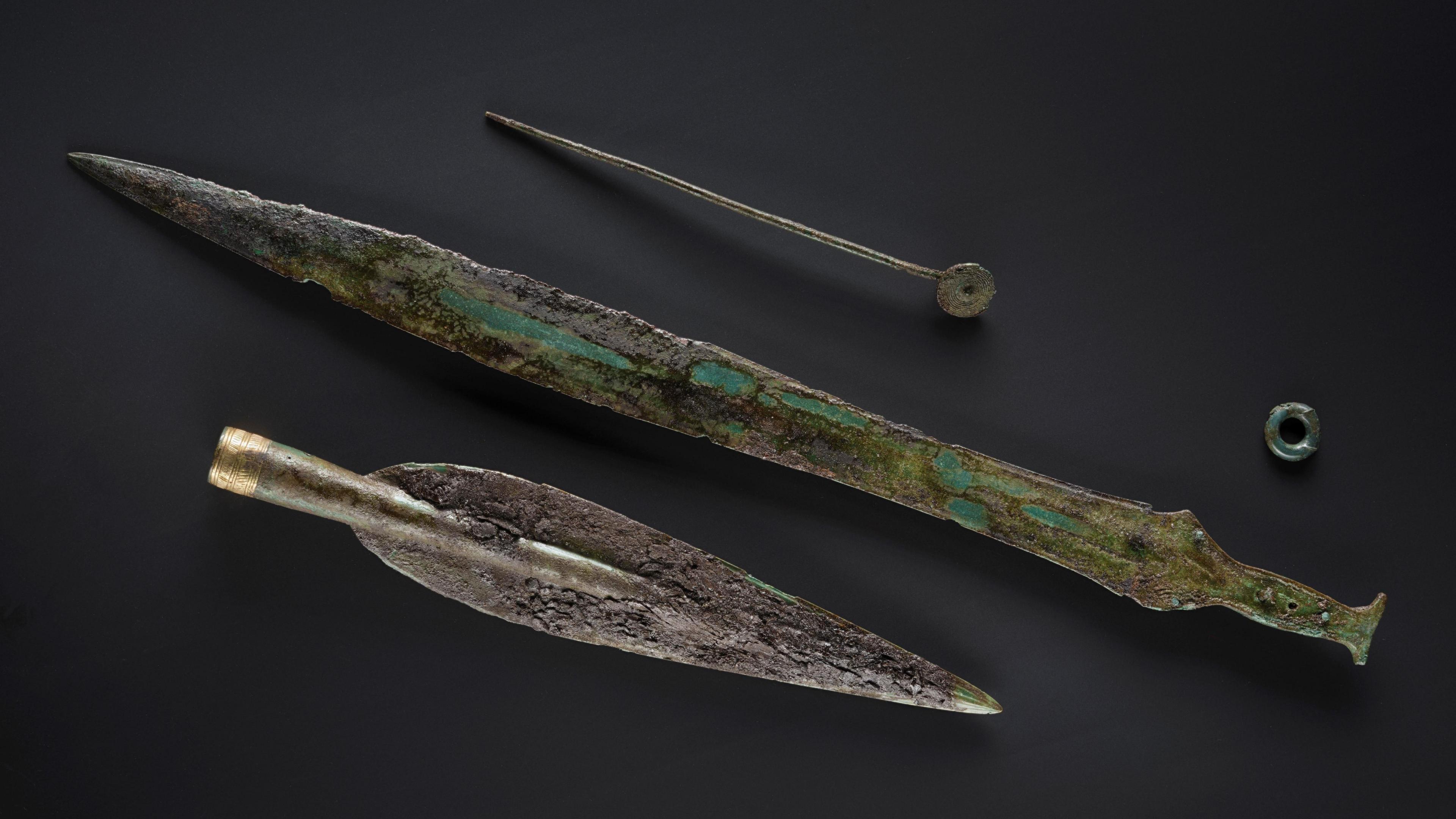 A bronze age spearhead and sword laid flat on a black surface.
