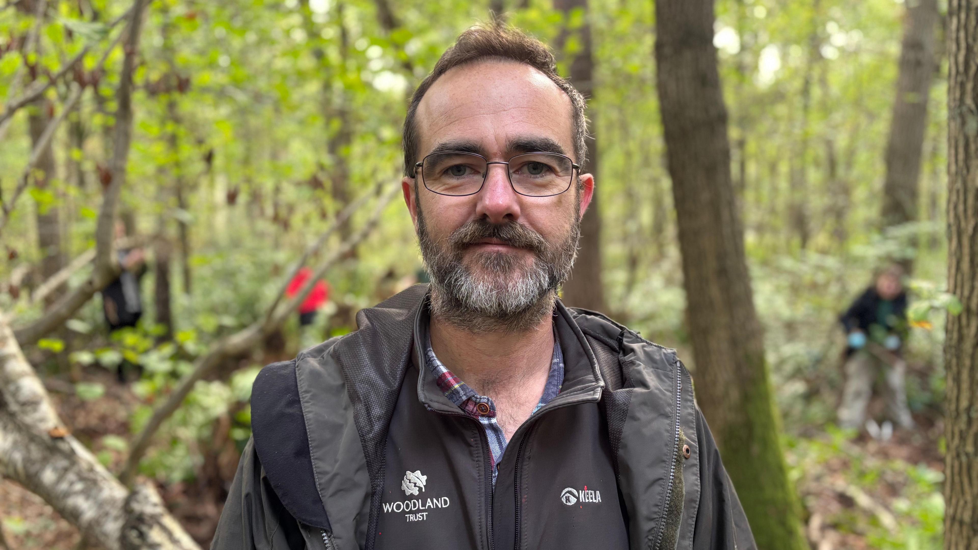 Woodland Trust site manager David Logan is pictured in the foreground wearing a black Woodland Trust fleece. Trees and people working are blurred in the background.