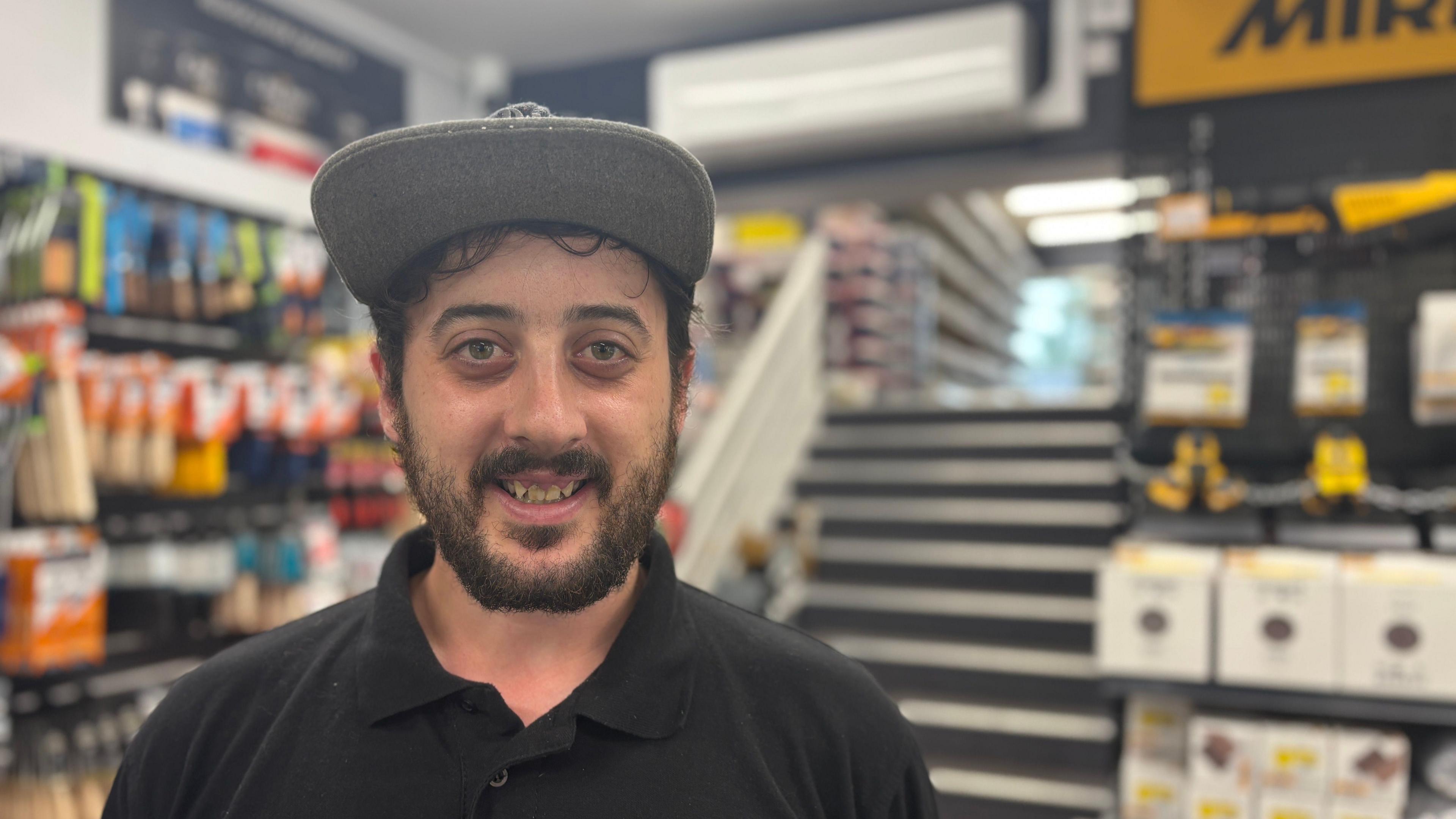 A brown haired man with a shirt brown beard smiles to the camera. He wears a black polo shirt and grey baseball cap. Behind him is a set of stairs as well as tools and paint brushes in the shop that he works in.