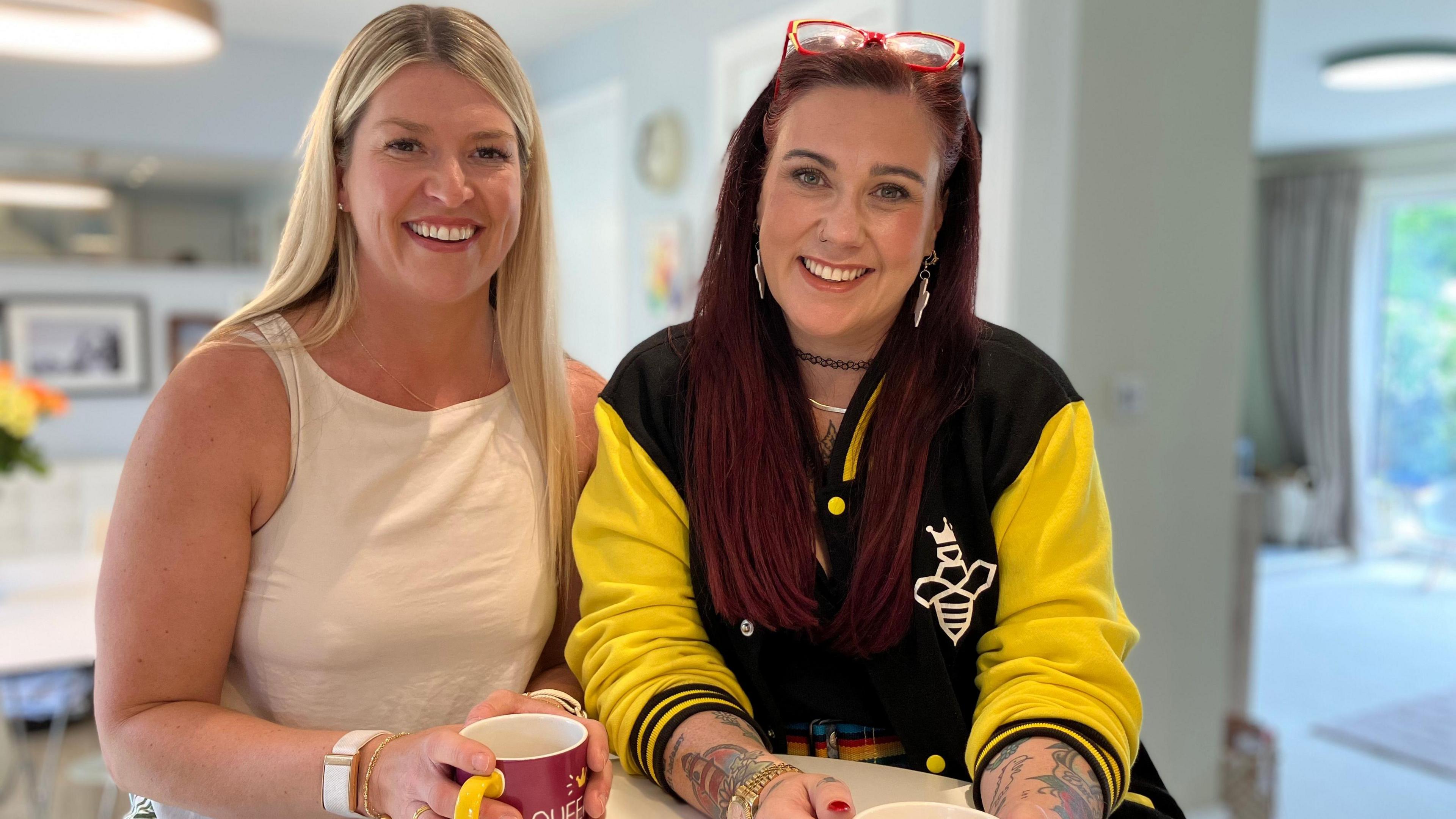 Emma Stirk and Dani Wallace smile at the camera and each have their hands cupped around mugs of tea 