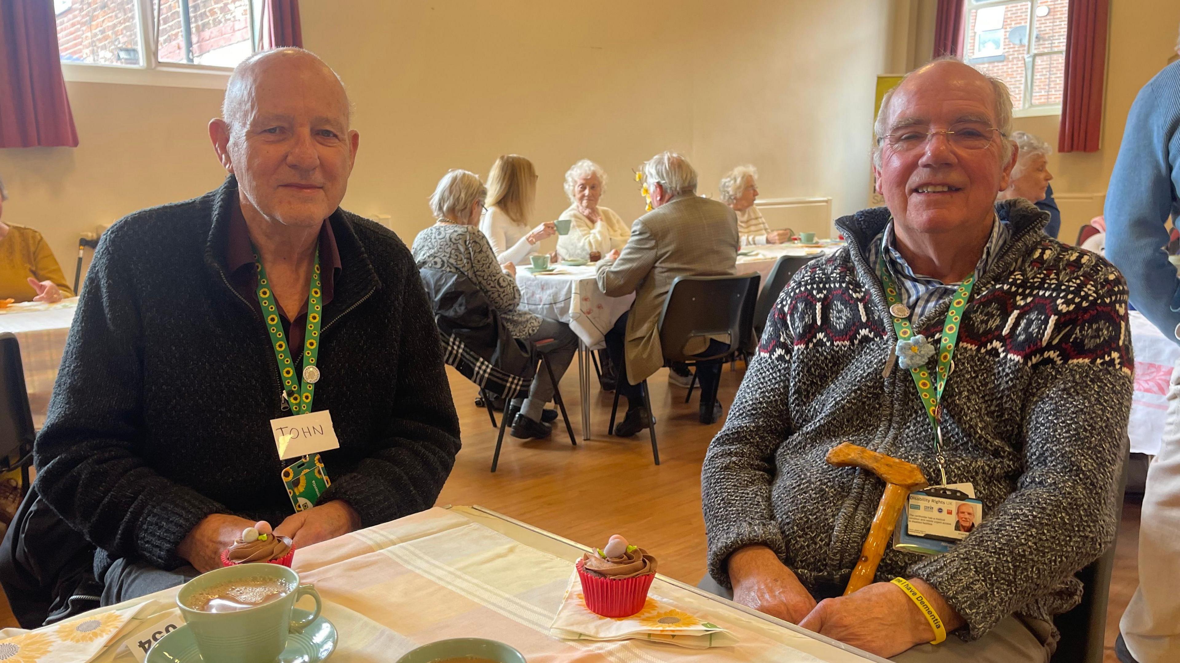 Two men sat at a table with cups of tea and cake. There is a table of elderly women behind them.