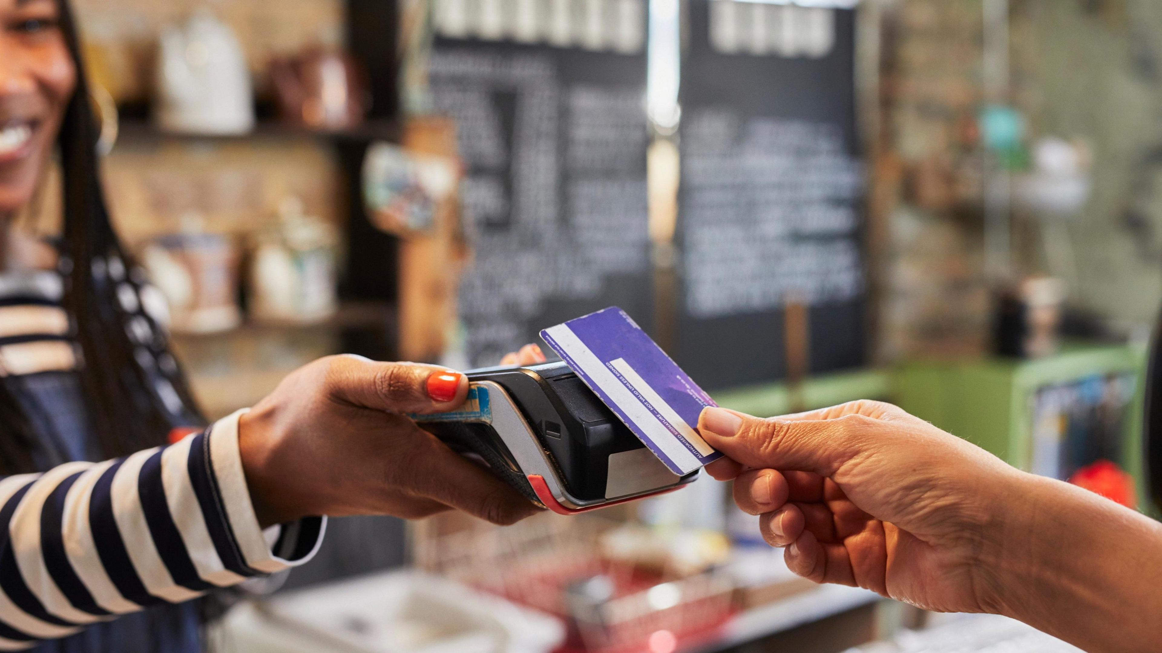 The hand of a customer putting their bank card on a card reader, being held by a shopkeeper.