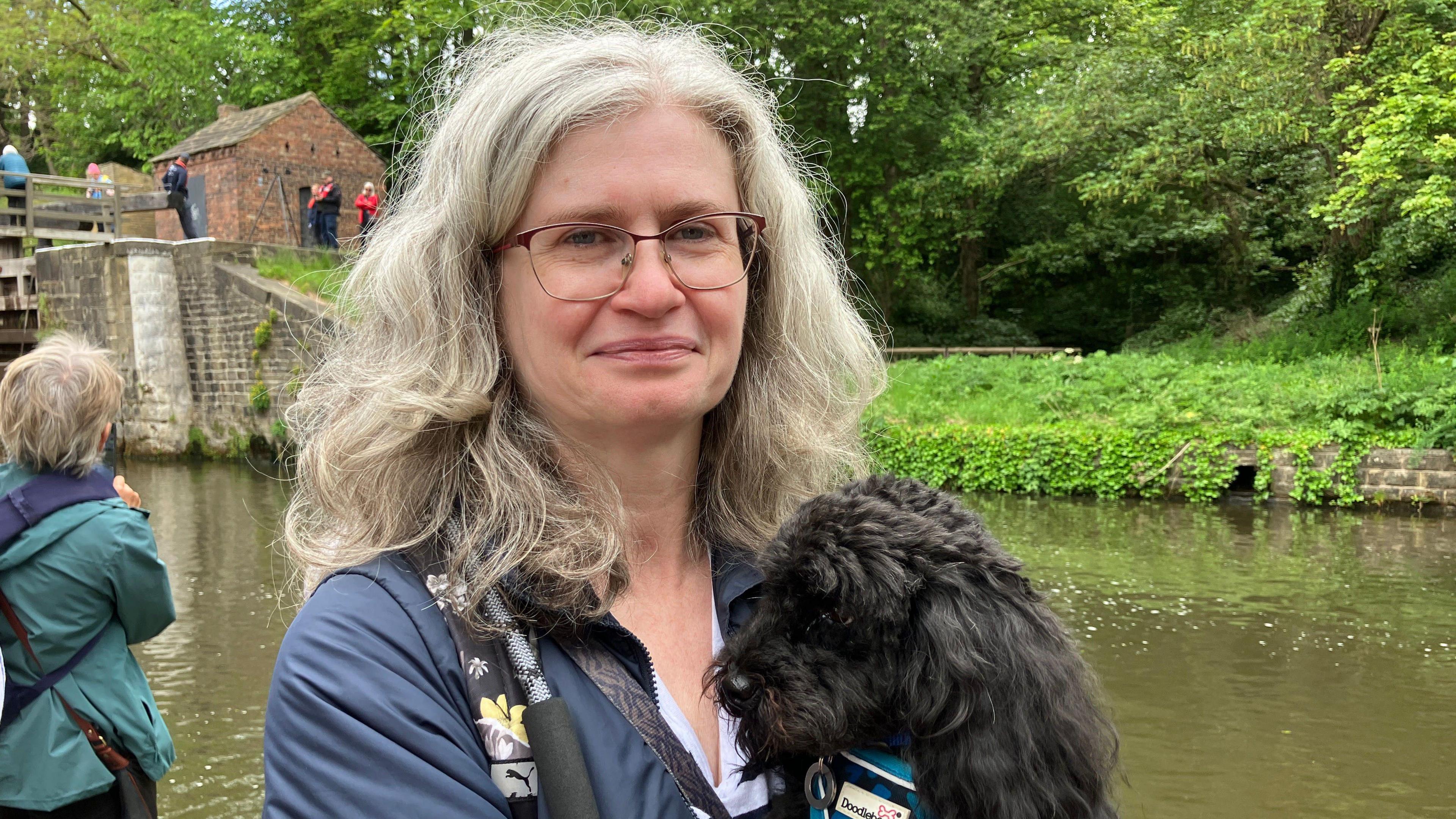 A woman stands in front of a canal. She wears glasses and a blue coat and holds a medium-sized black dog in her arms.