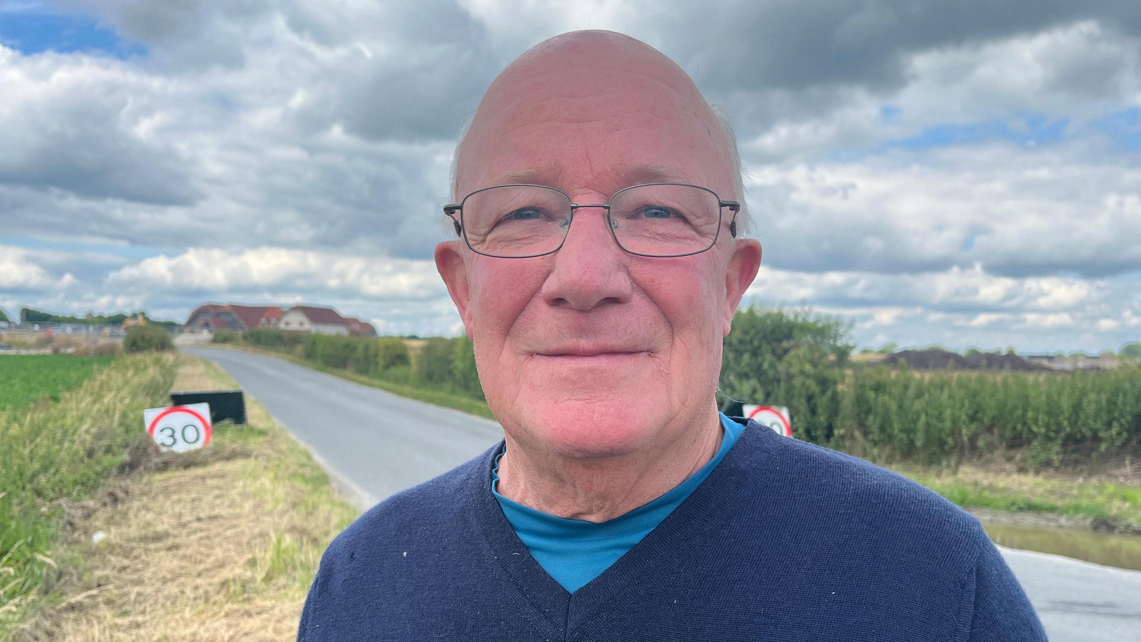 A man in a blue v-necked jumper over a lighter blue t-shirt wearing glasses and standing on the side of a road