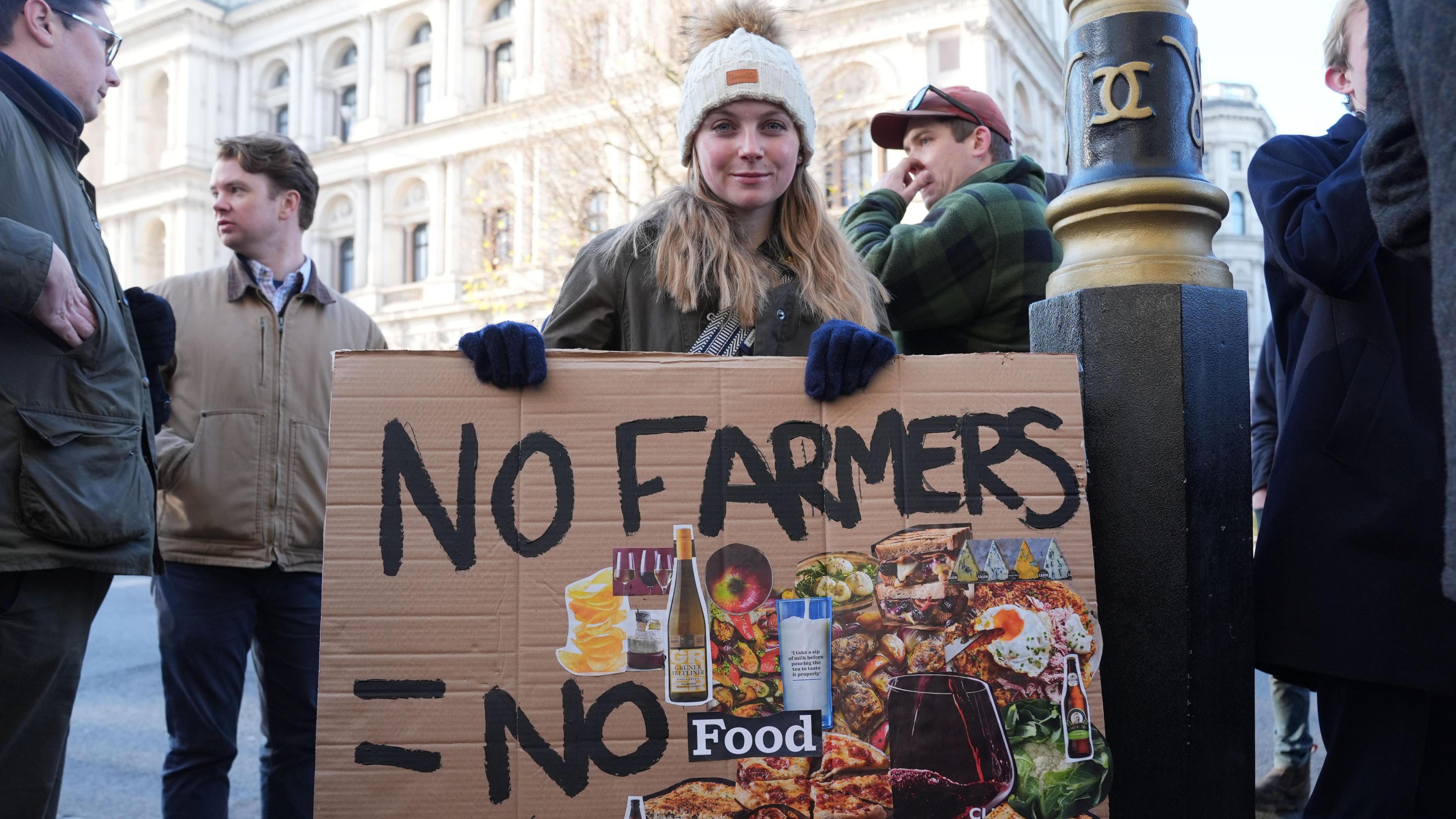 Young woman with long blond hair and wearing a bobble hat holding a banner outside parliament which reads no farmers + no food
