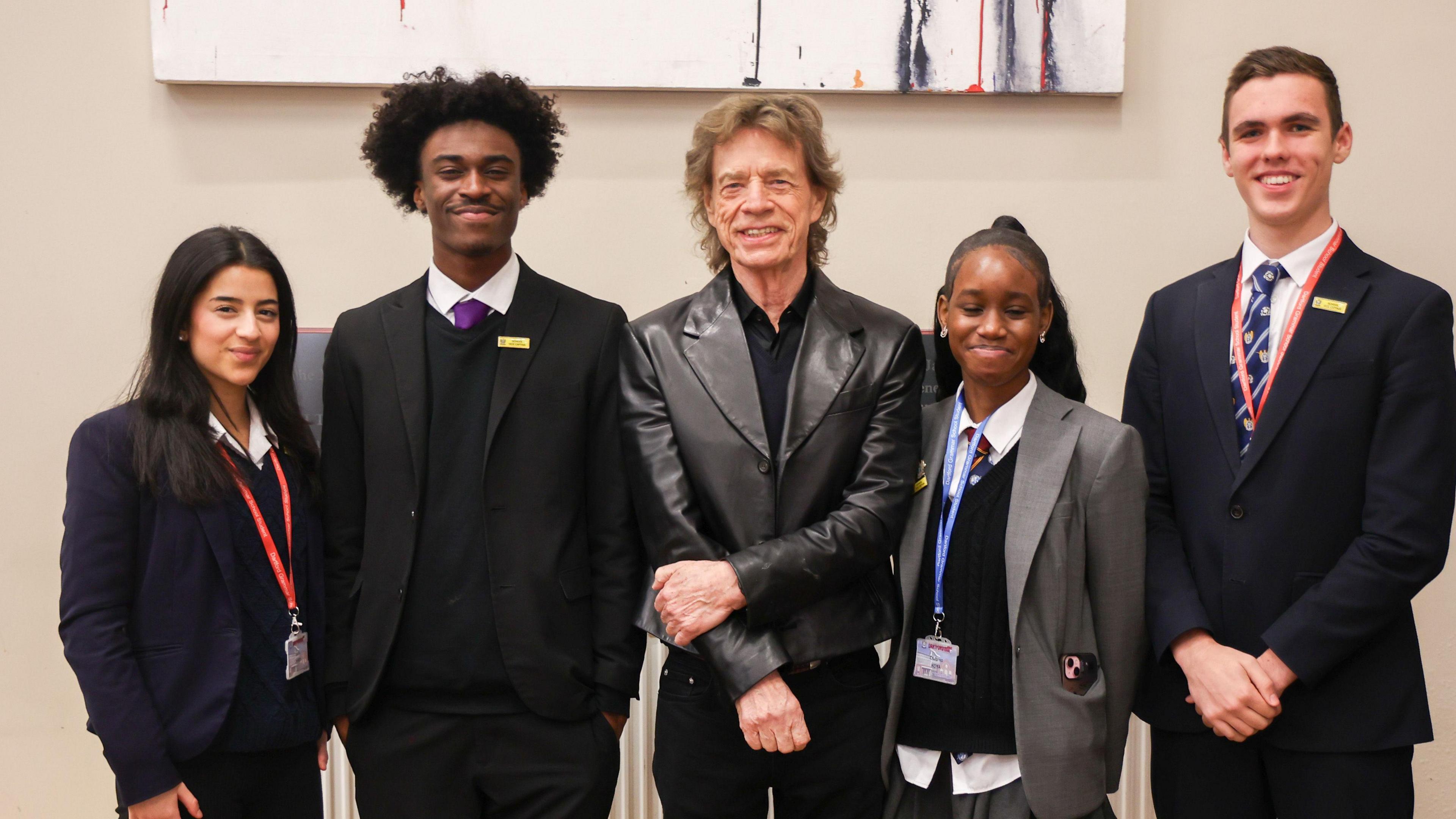 Sir Mick Jagger wearing a dark shirt and leather jacket. Four older children wearing school uniform and lanyards are stood either side of him. All are smiling at the camera.