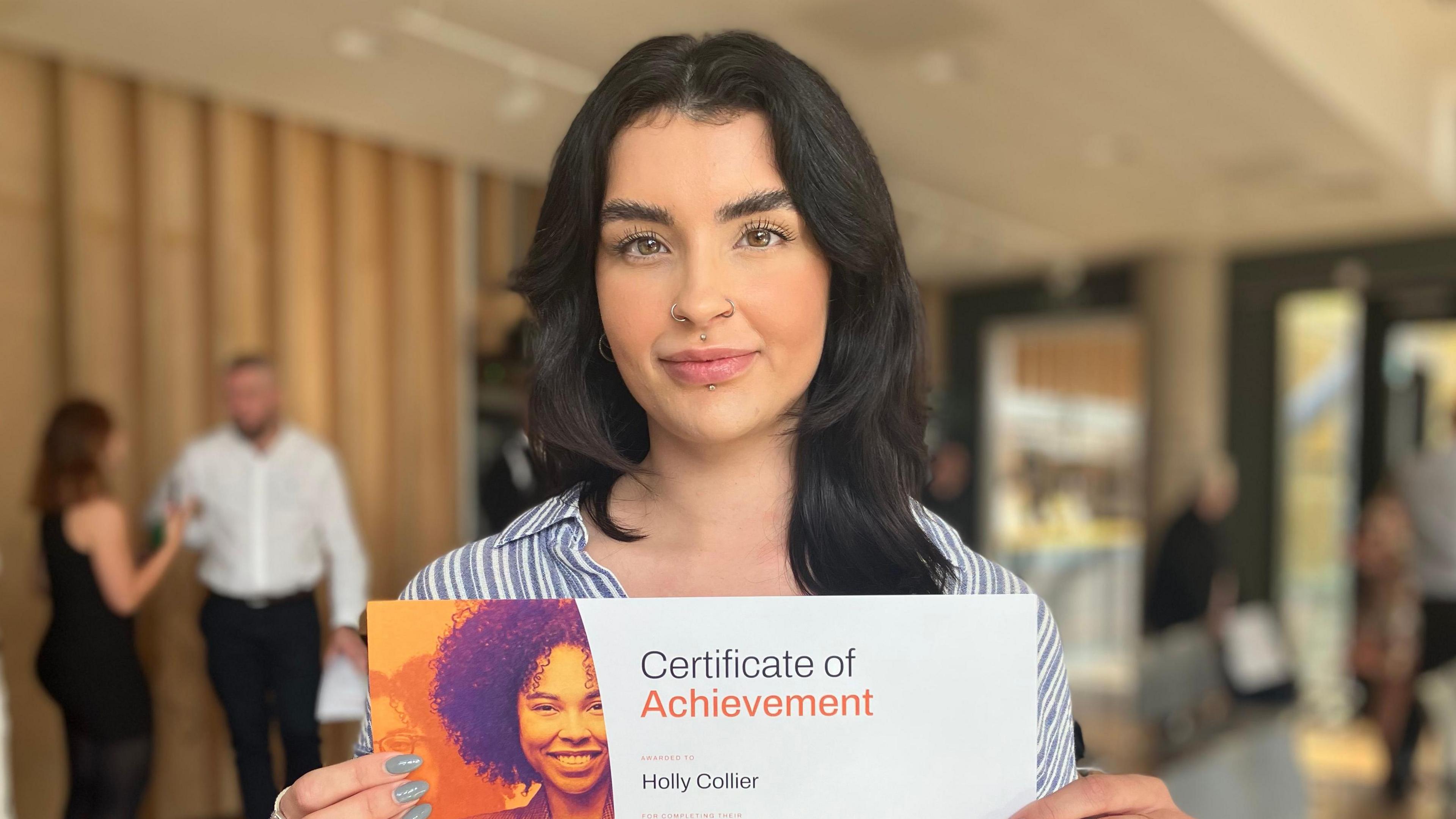 Holly Collier holding up a certificate, wearing a striped shirt in a conference room.