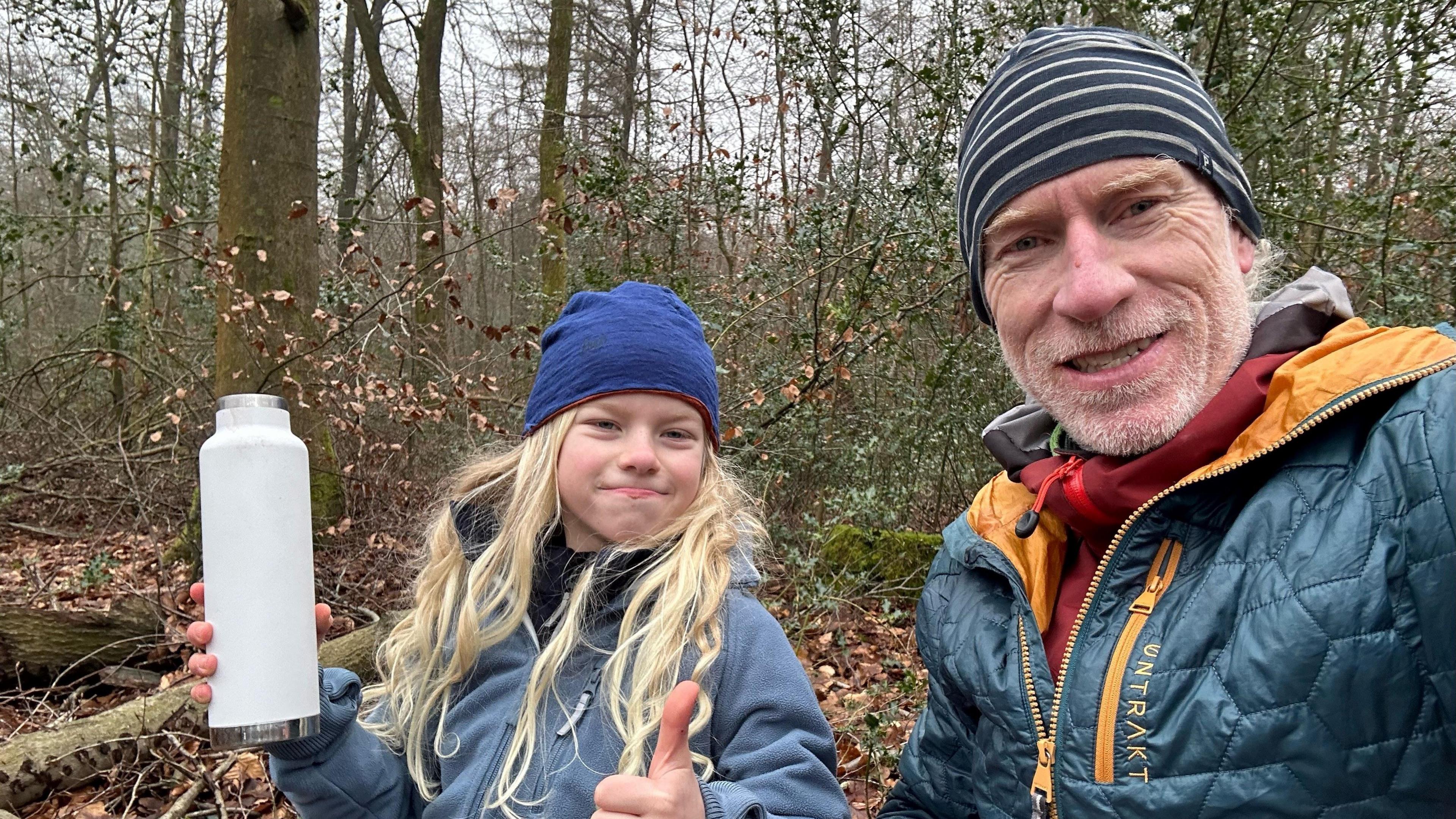 Two people sat on a log in the forest. Orran, pictured left, is wearing a blue hat, a light blue fleece and is holding a light blue water bottle. David, pictured right, is wearing a stripy hat and a teal blue and yellow coat 