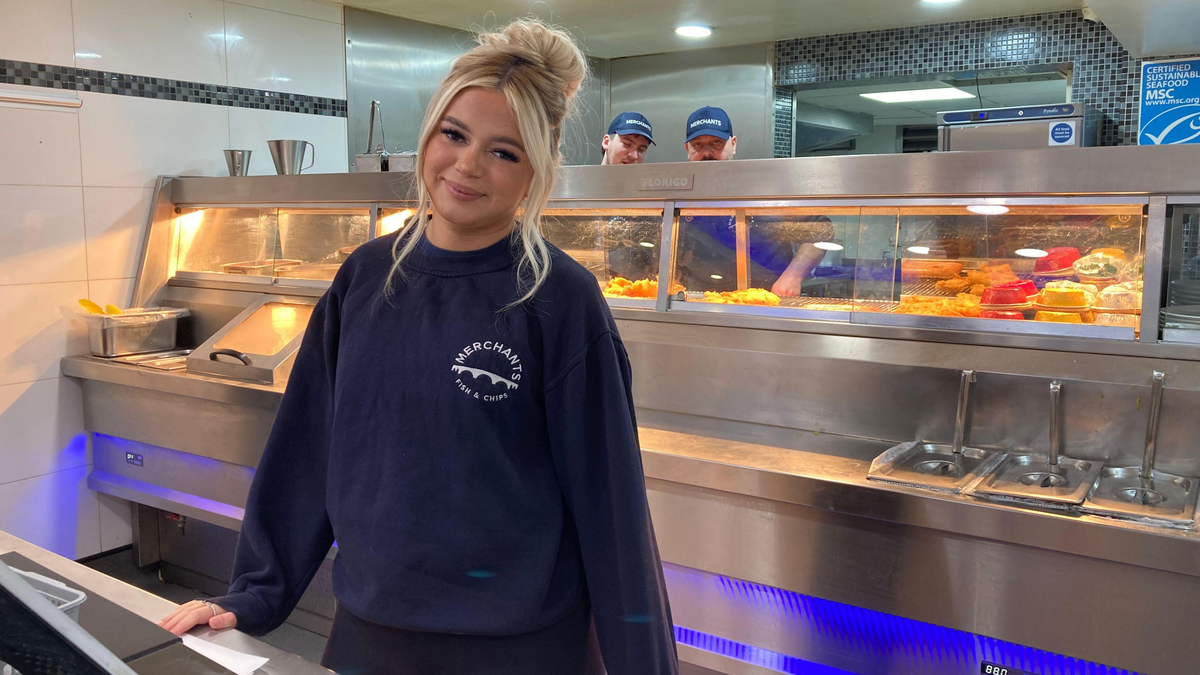 Destiny Harold posing for a photo. She has blonde hair and is behind the counter of a chip shop. There are two men behind her preparing fried food. 