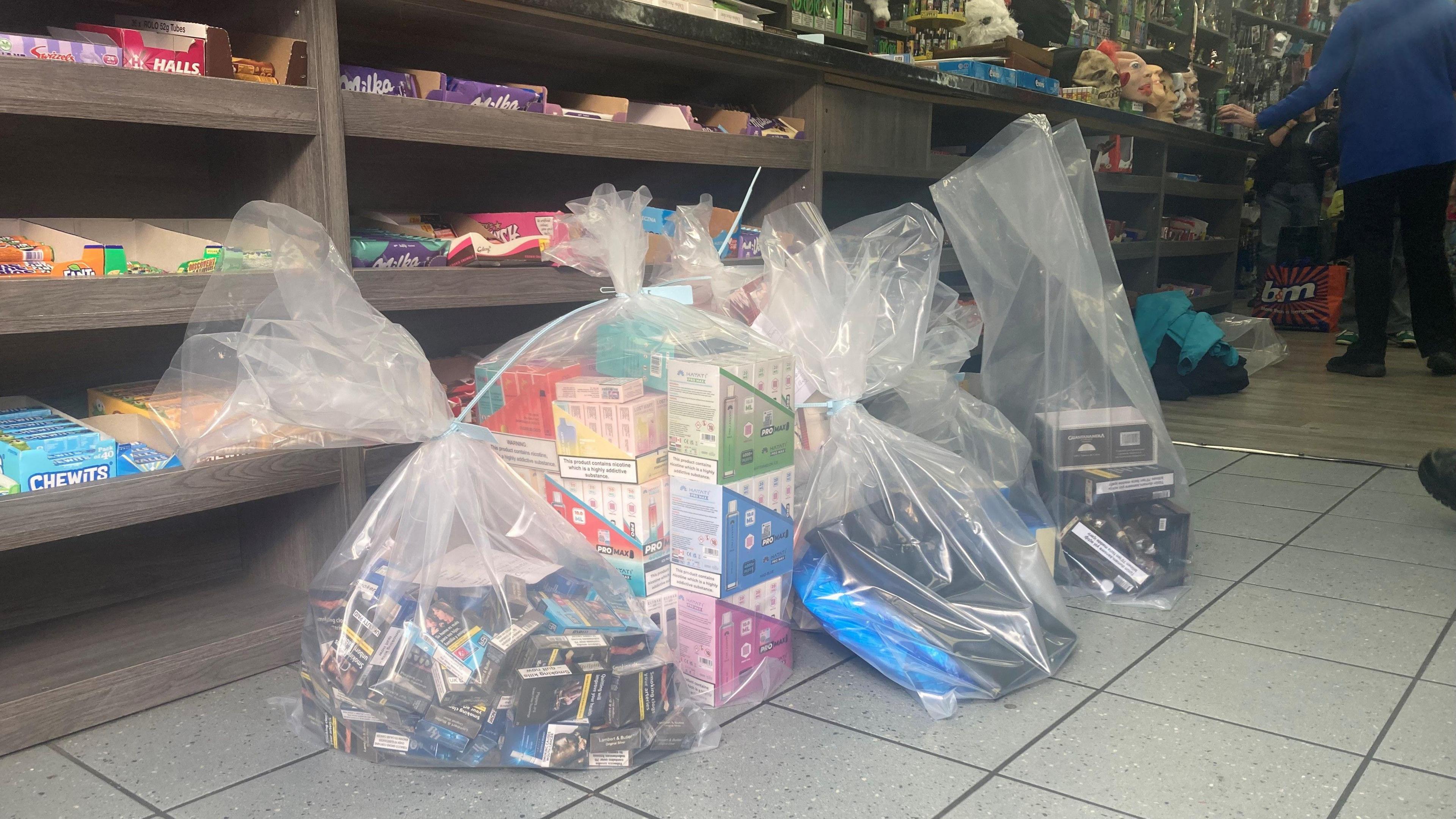 A shop floor with shelves in the background and three large plastic bags with seized cigarettes and vapes inside.