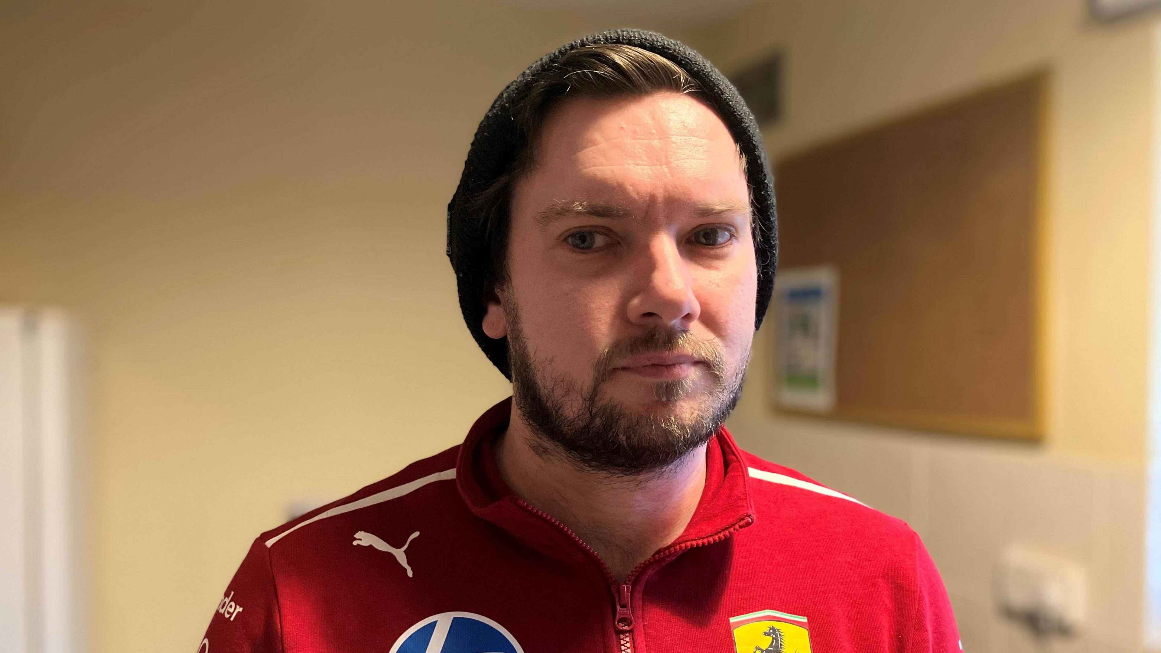 Andrew stares at the camera. He wears a red top with sports logos on it. A has a short beard and wears a black hat. The background is beige walls with a noticeboard to the right. 