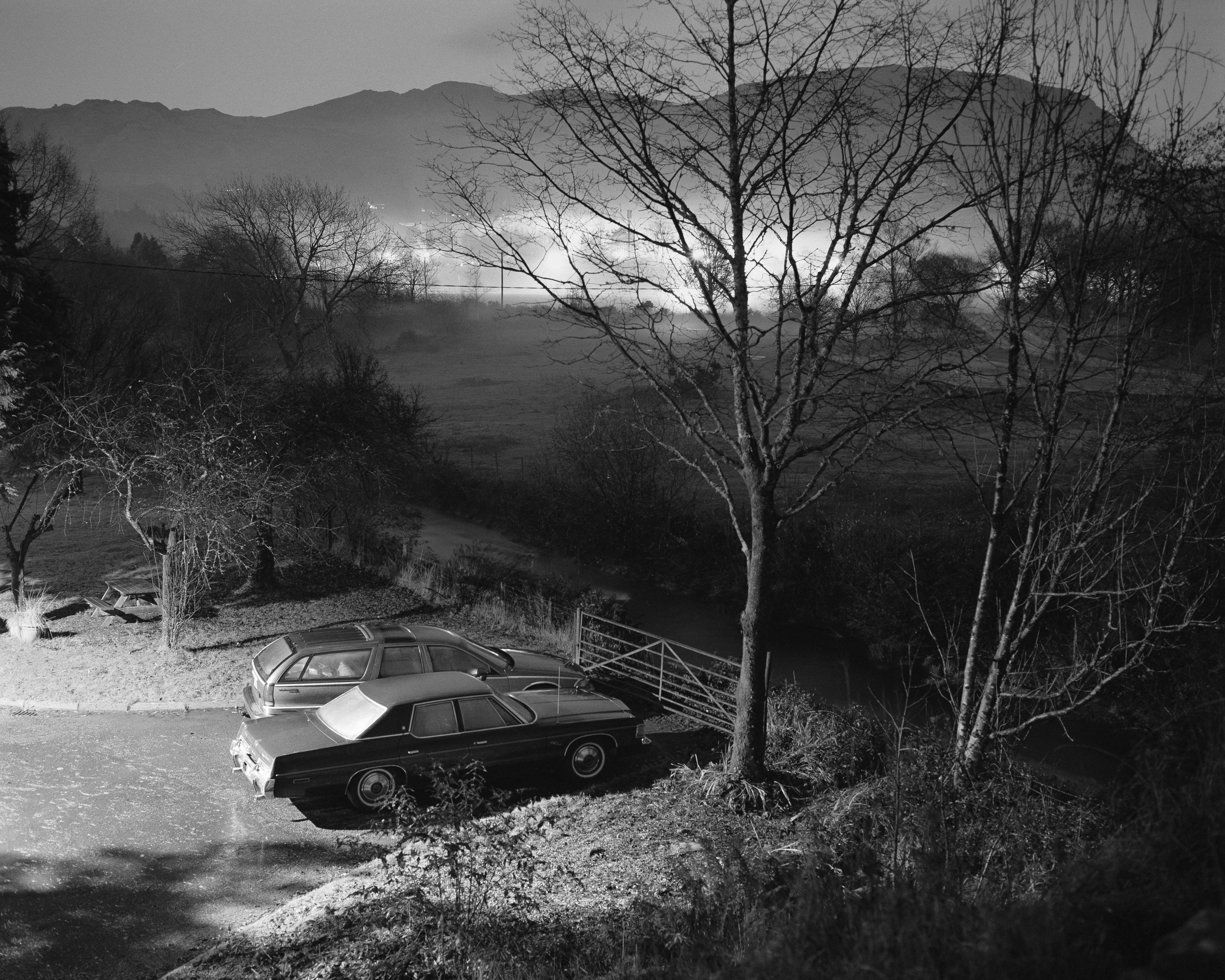 Kyle's black and white photograph of two cars parked by a metal gate and care trees with misty hills behind them.