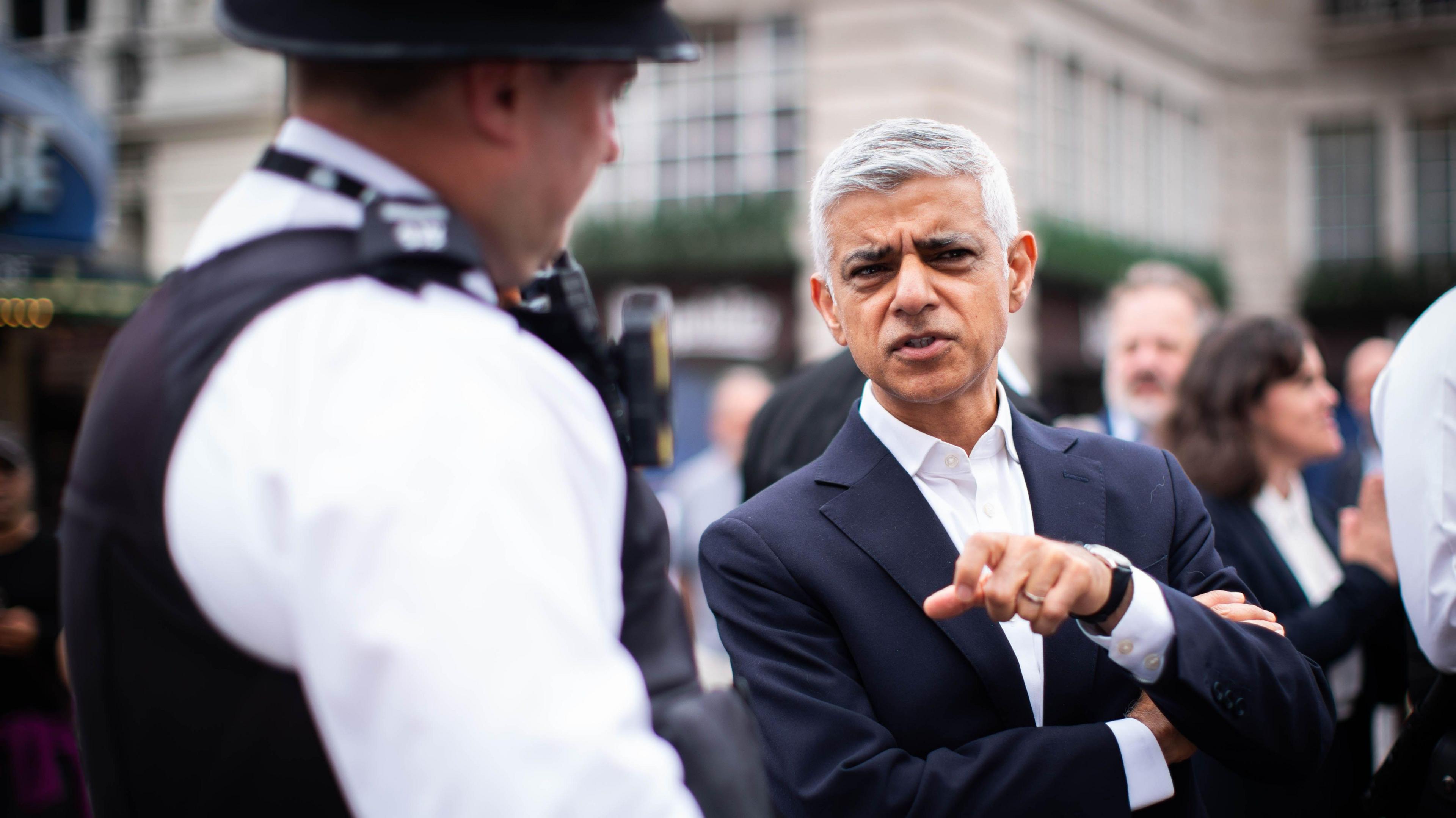 Sir Sadiq Khan is seen in a dark suit speaking to a Metropolitan Police officer wearing a uniform and helmet in a busy street.