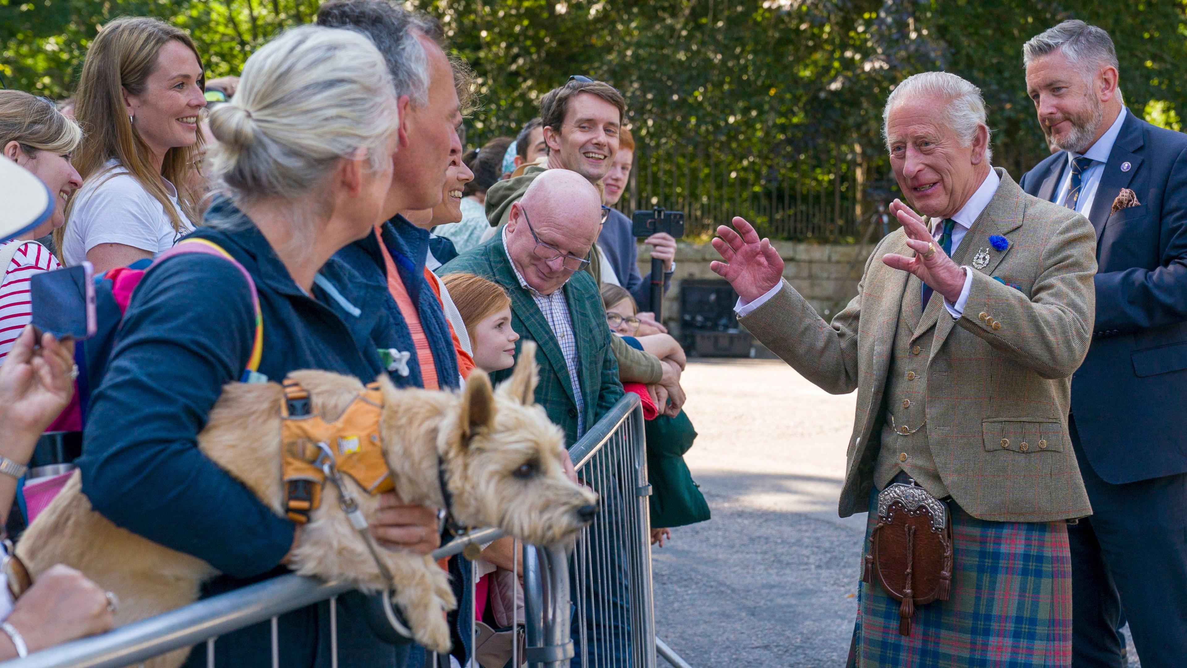 King Charles III meets members of the public after carrying out an inspection of the Balaklava Company, 5th Battalion, The Royal Regiment of Scotland, at the gates of Balmoral. He is smiling and holding his hands in front of him while talking to members of the public who are standing behind a metal barrier.