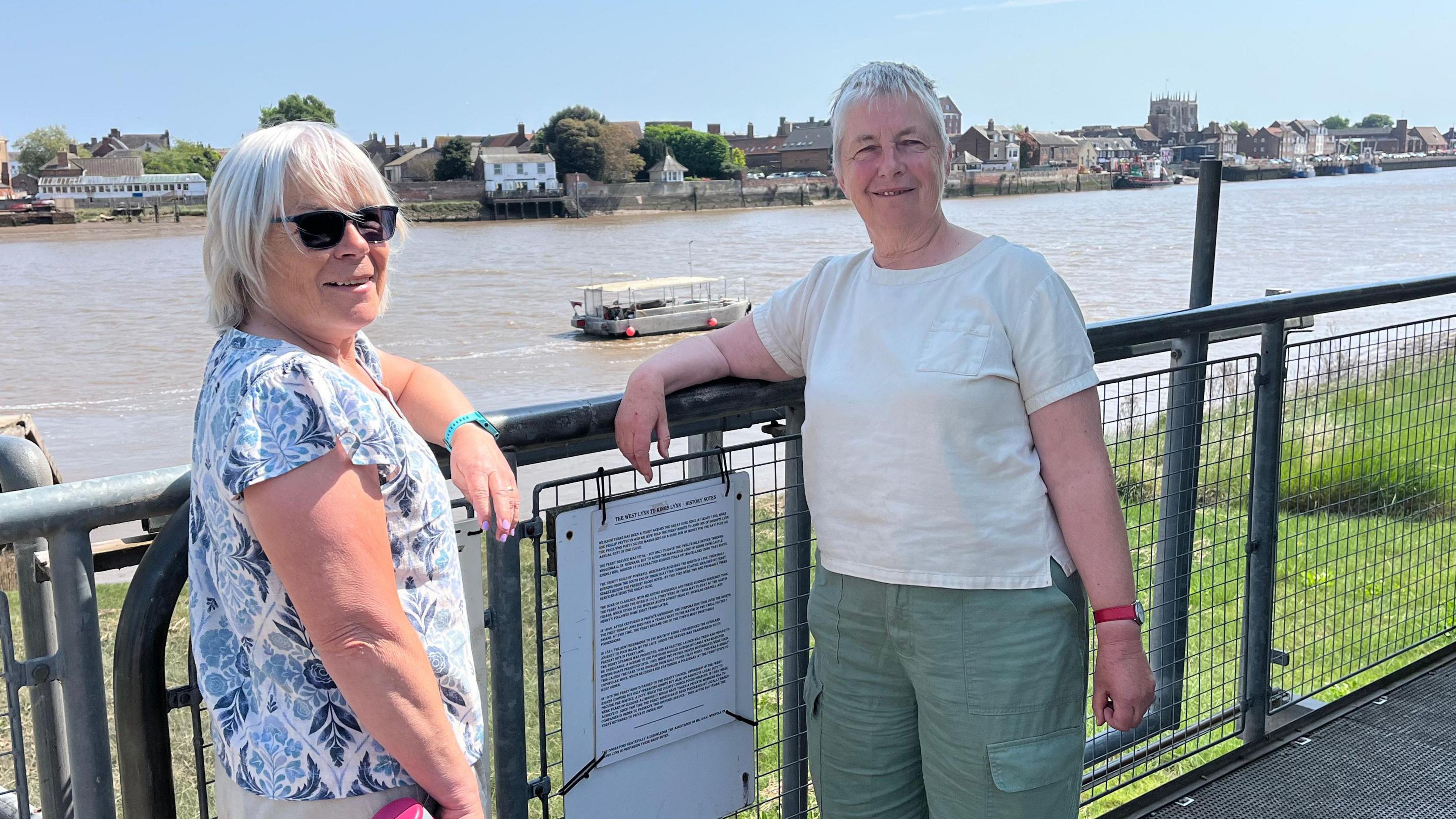 Alison Fletcher and Karen Champion stand in front of the Ferry which is moored in The Great Ouse river.
Alison is wearing a white and blue top and dark sunglasses and Karen is wearing a white top and green shorts.