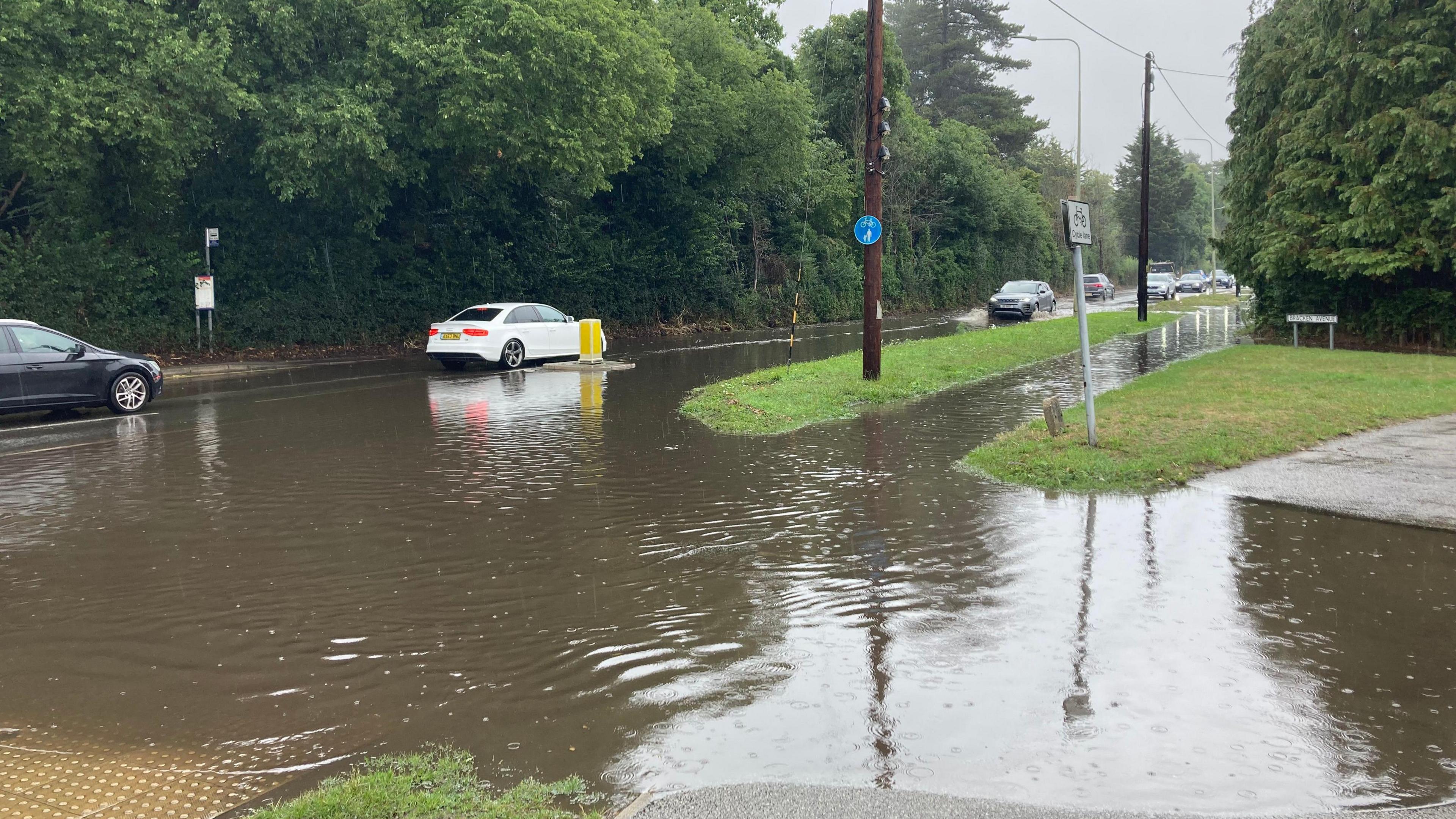 A road is pictured covered in flood water. Drivers attempt to drive through the water.
