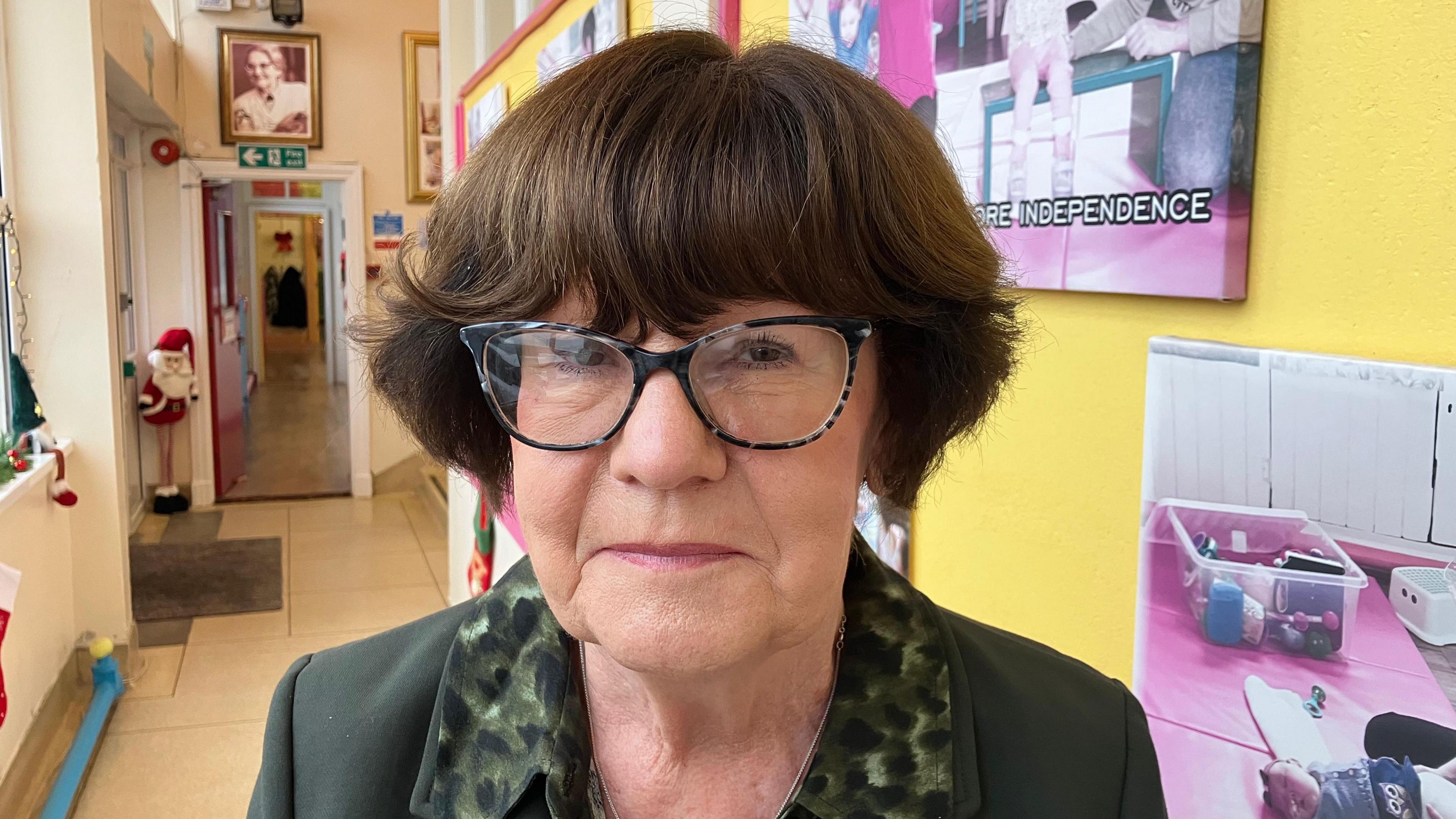 A woman with short brown hair and dark rimmed glasses is looking into the camera. She is wearing a green patterned shirt with a dark green jacket. She is standing in a yellow school corridor with a number of pictures on the walls. 