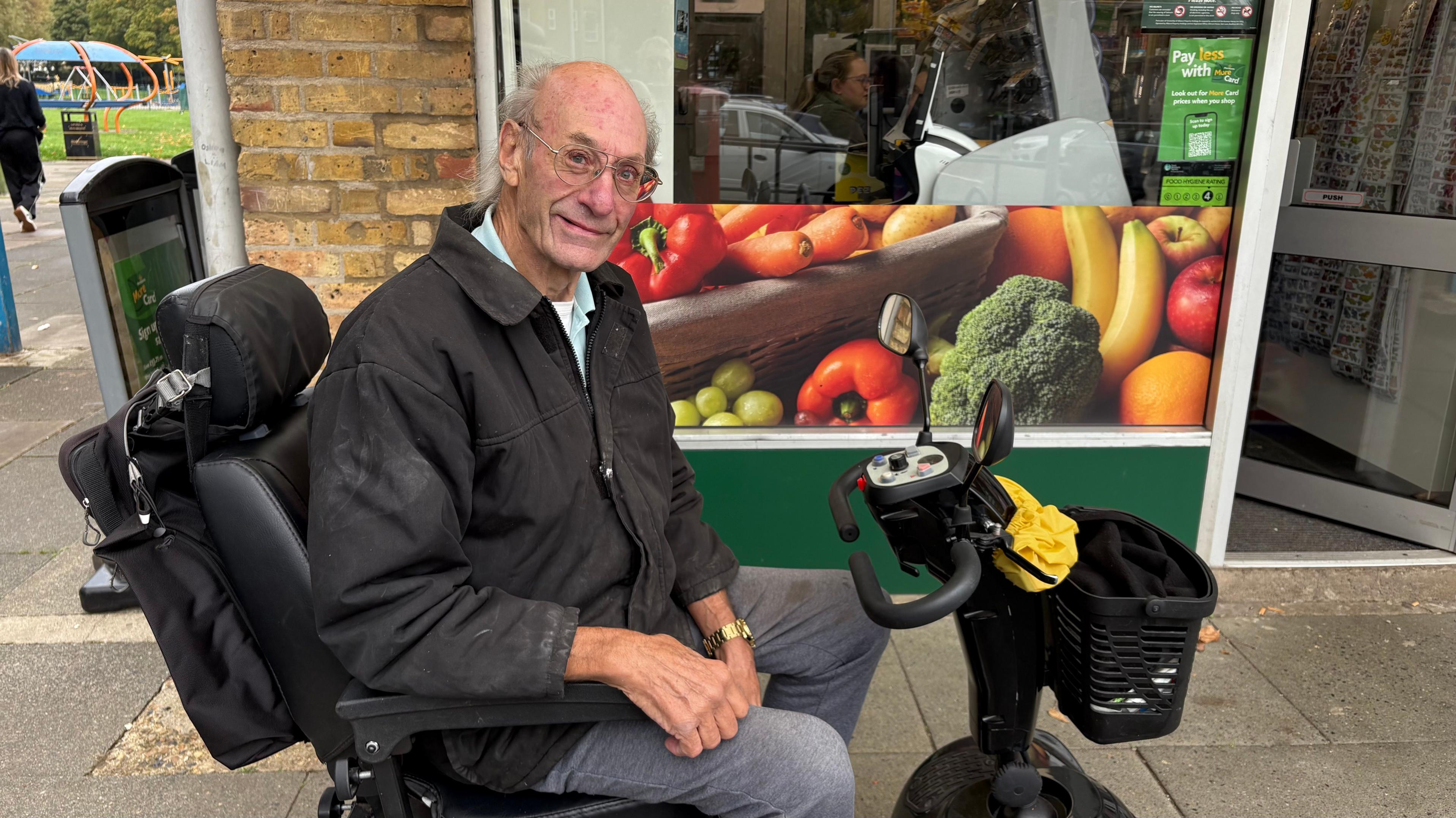 Will Maddox with short white hair and glasses, smiling at the camera while wearing a black zip-up jacket and grey trousers. He is sitting on a black electric scooter with a shopping bag tied over the handlebars. There is a shop behind him with a poster advertising fruit and vegetables.