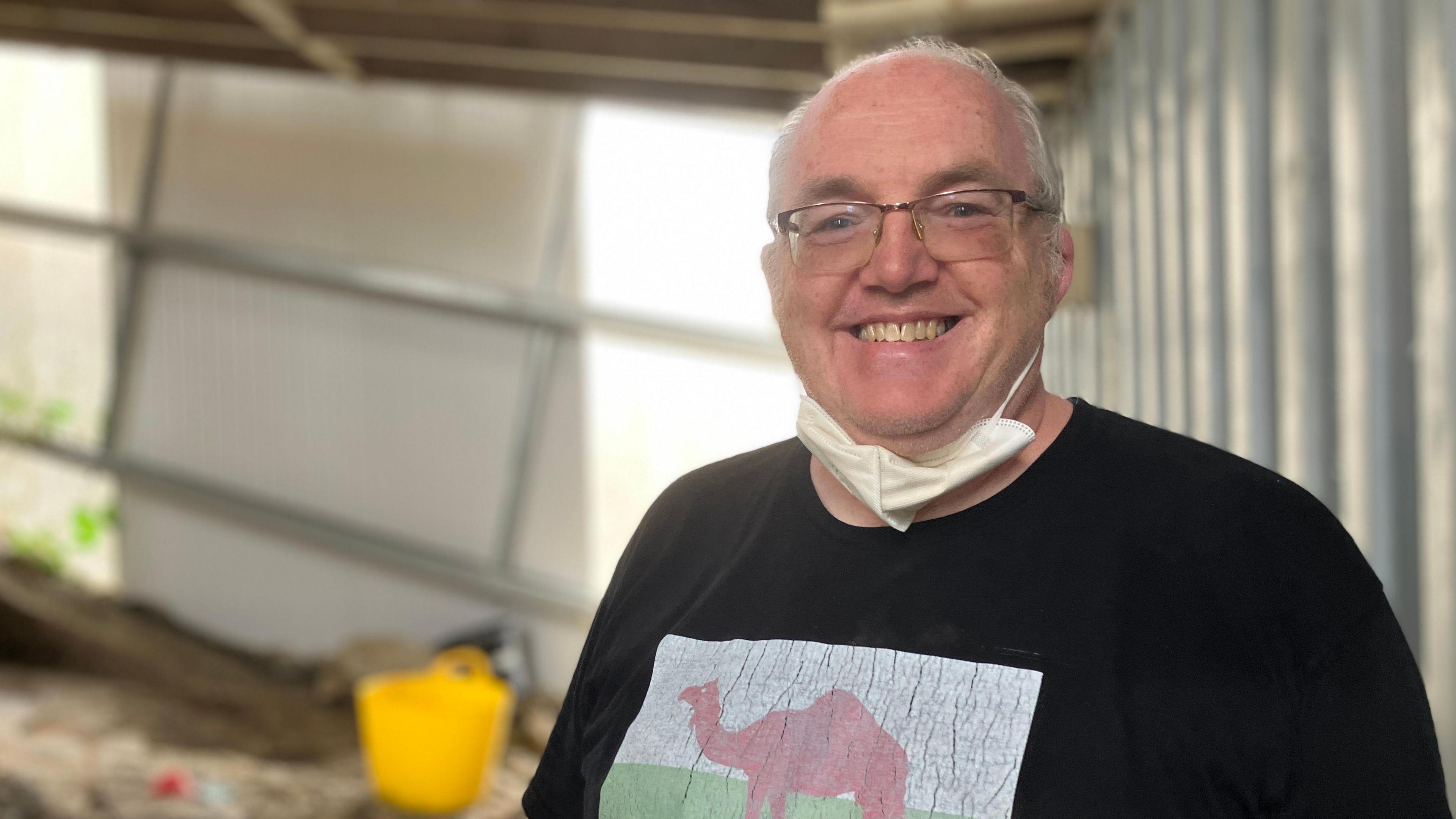 Nic Jones photographed at the site, he wears a mouth mask tucked under his chin. He has blue eyes, silver hair on the sides and wears square reading glasses. He smiles showing his teeth and wears a black t-shirt. 