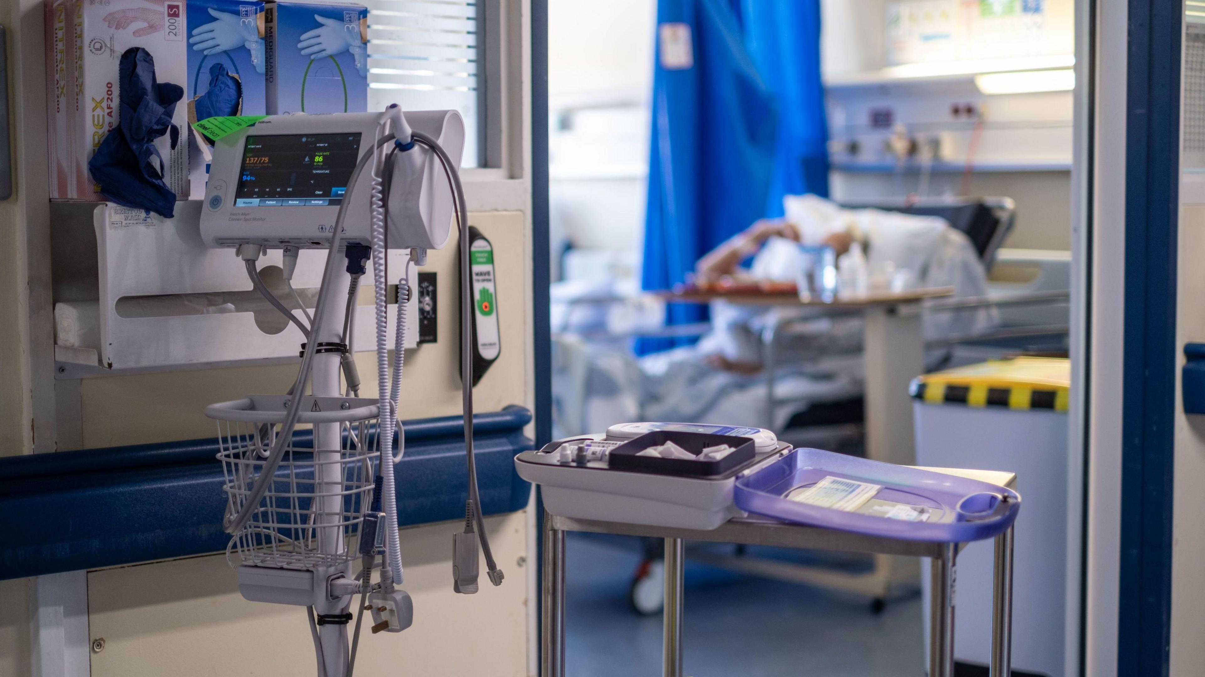 General view of medical equipment on a NHS hospital ward. There is equipment at the front of the picture, including a monitor and packets of gloves, and a patient can be seen in a hospital bed in the background, next to a blue curtain.