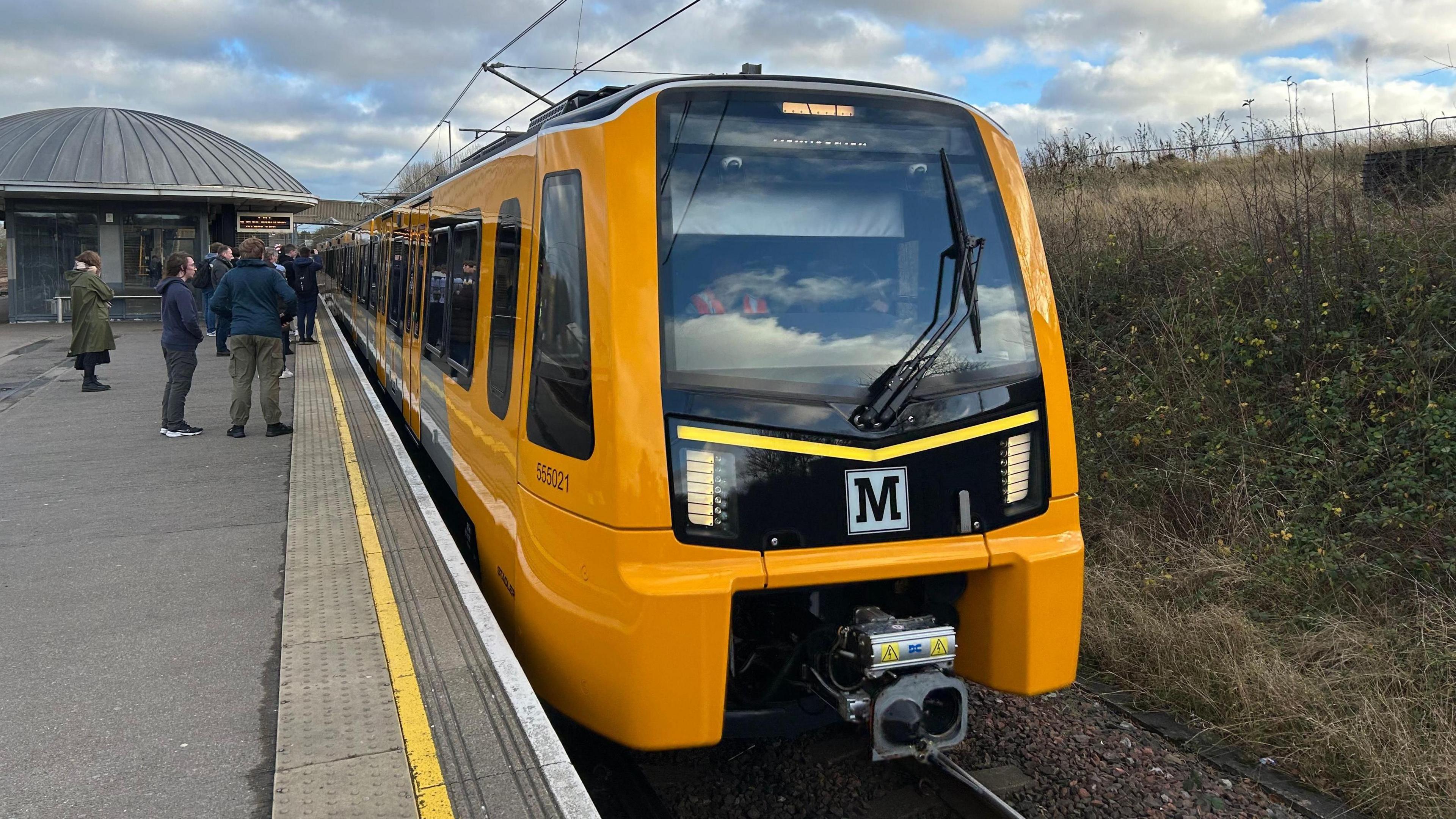 The first new Tyne and Wear Metro train in passenger service. The train is yellow and has the Metro M logo on its front. It is pulled into a station and passengers are waiting to board.