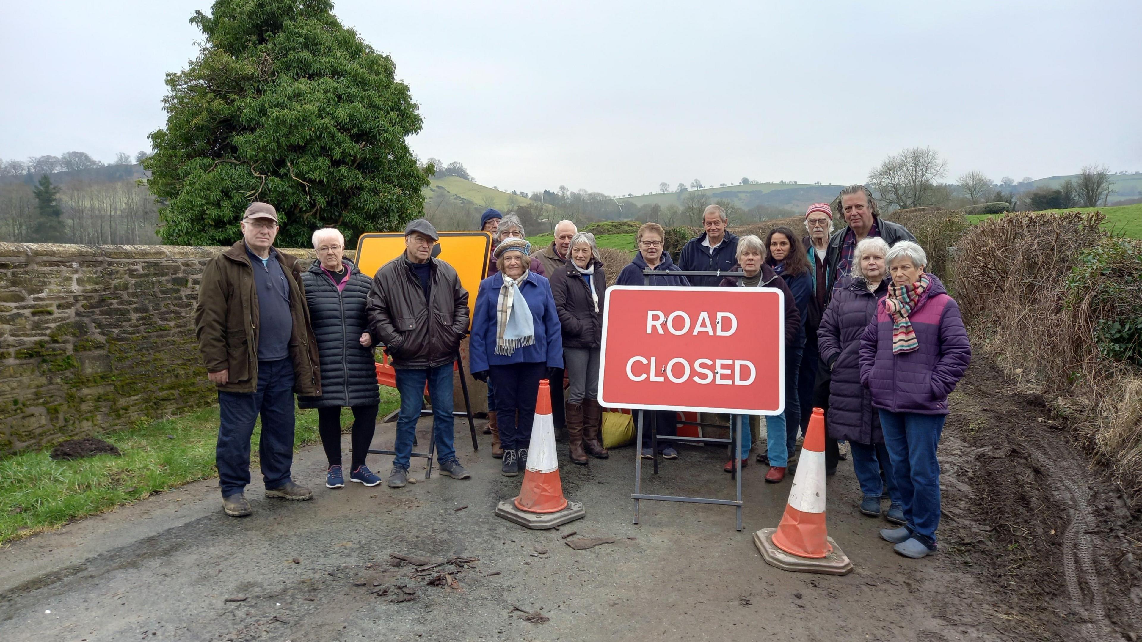 A group of people standing on a rural road behind a road closed sign next to two traffic cones. Trees, hills and fields can be seen in the background.