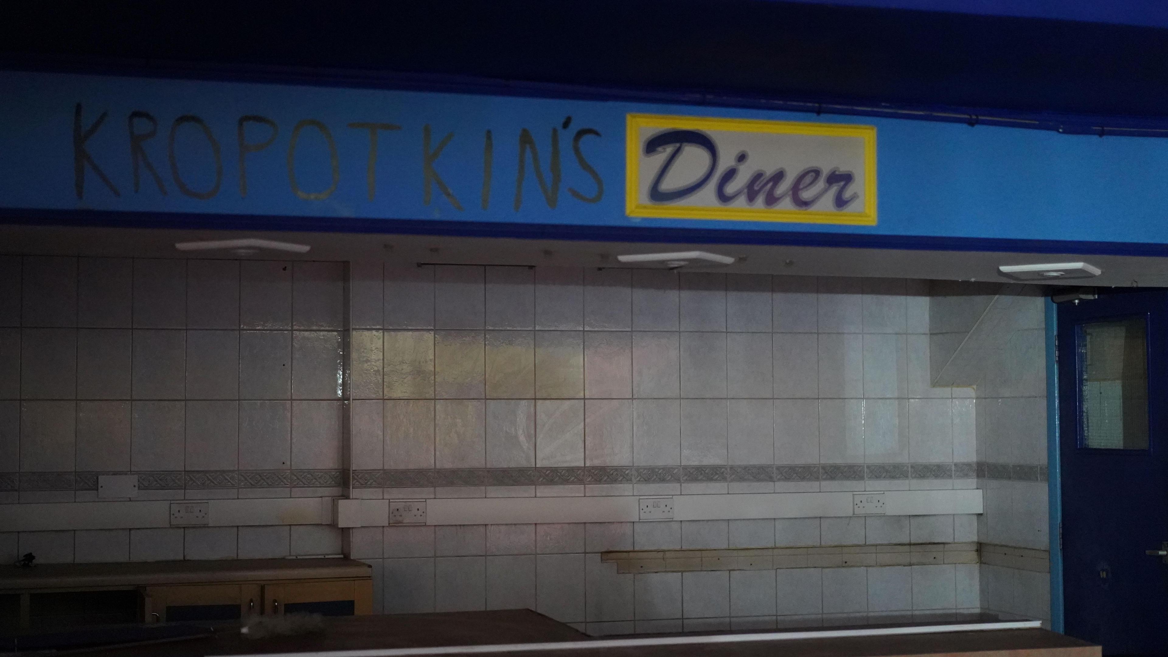 A sign for a diner, behind it are white-tiled walls. Someone has written "Kropotkin's" in black ink before the word "diner".