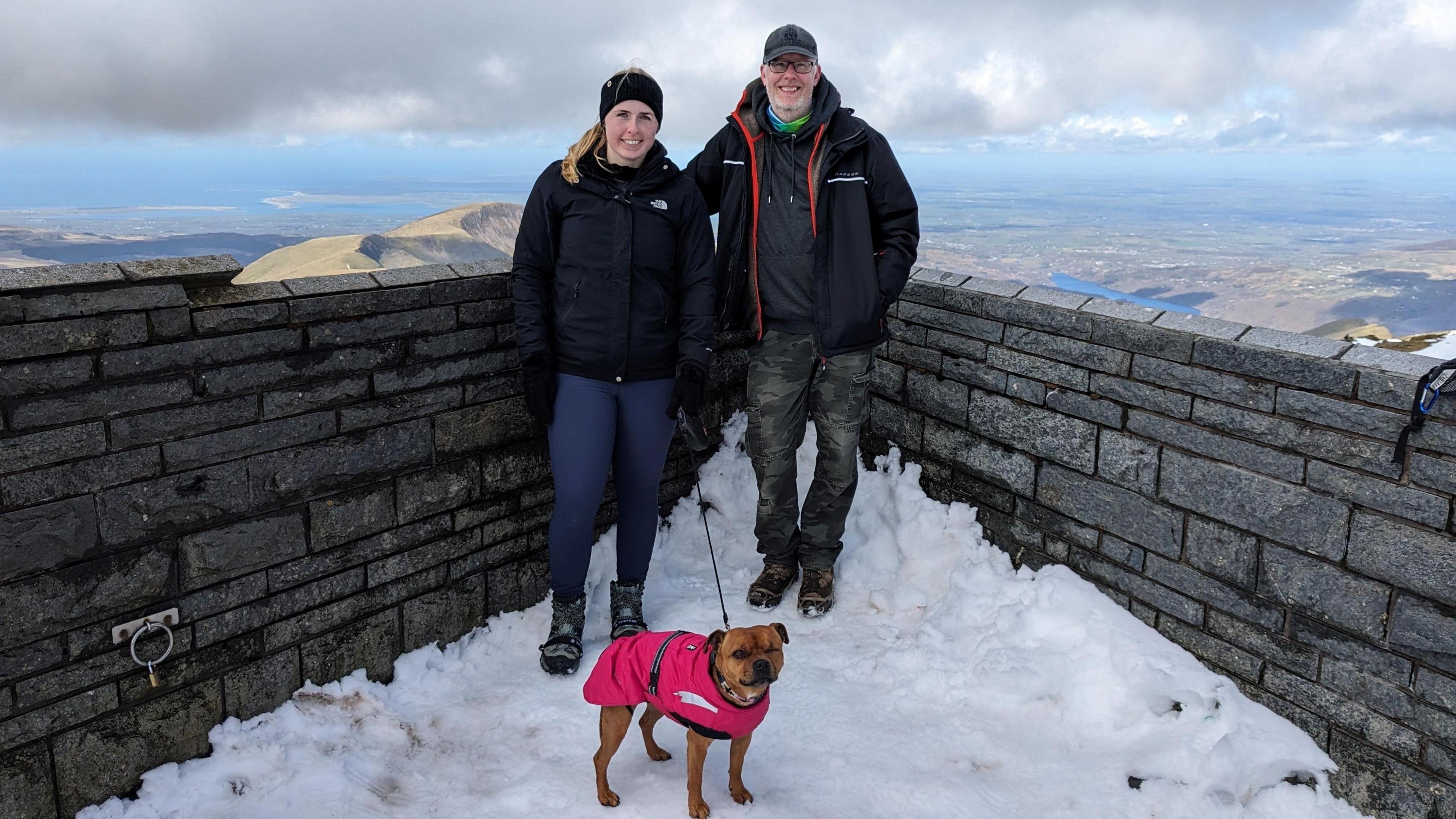 Jennie and Stuart on a snowy ledge with a dog. There is a grey brick wall behind them and a mountainous landscape behind them. They are all dressed in extra layers, including the dog.