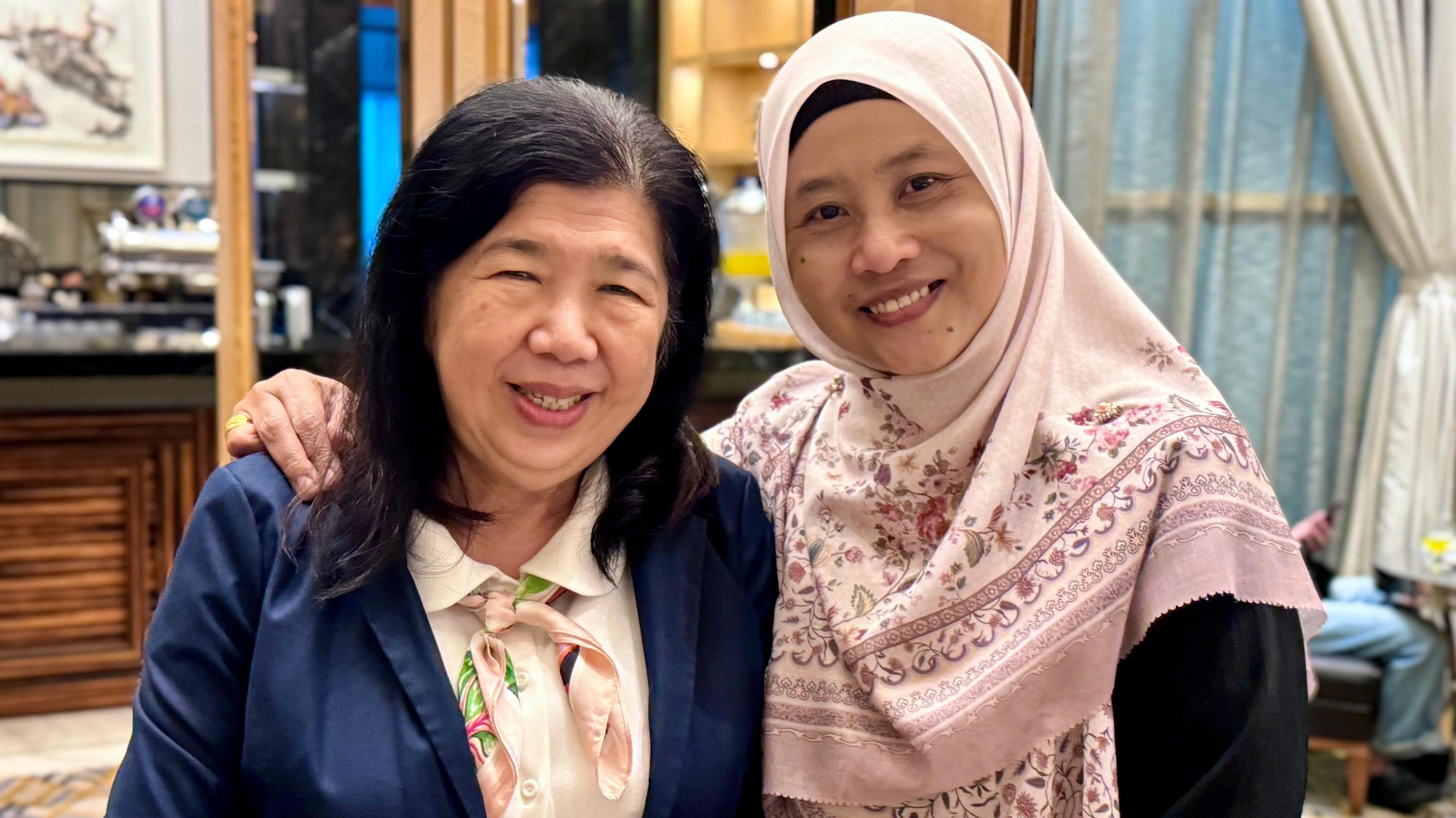 Susanna Liew wears a blue blazer, white shirt and pink neck scarf while Norhayati wears a pink patterned hijab. Both women are smiling and Norhayati's arm is around Susanna's shoulders