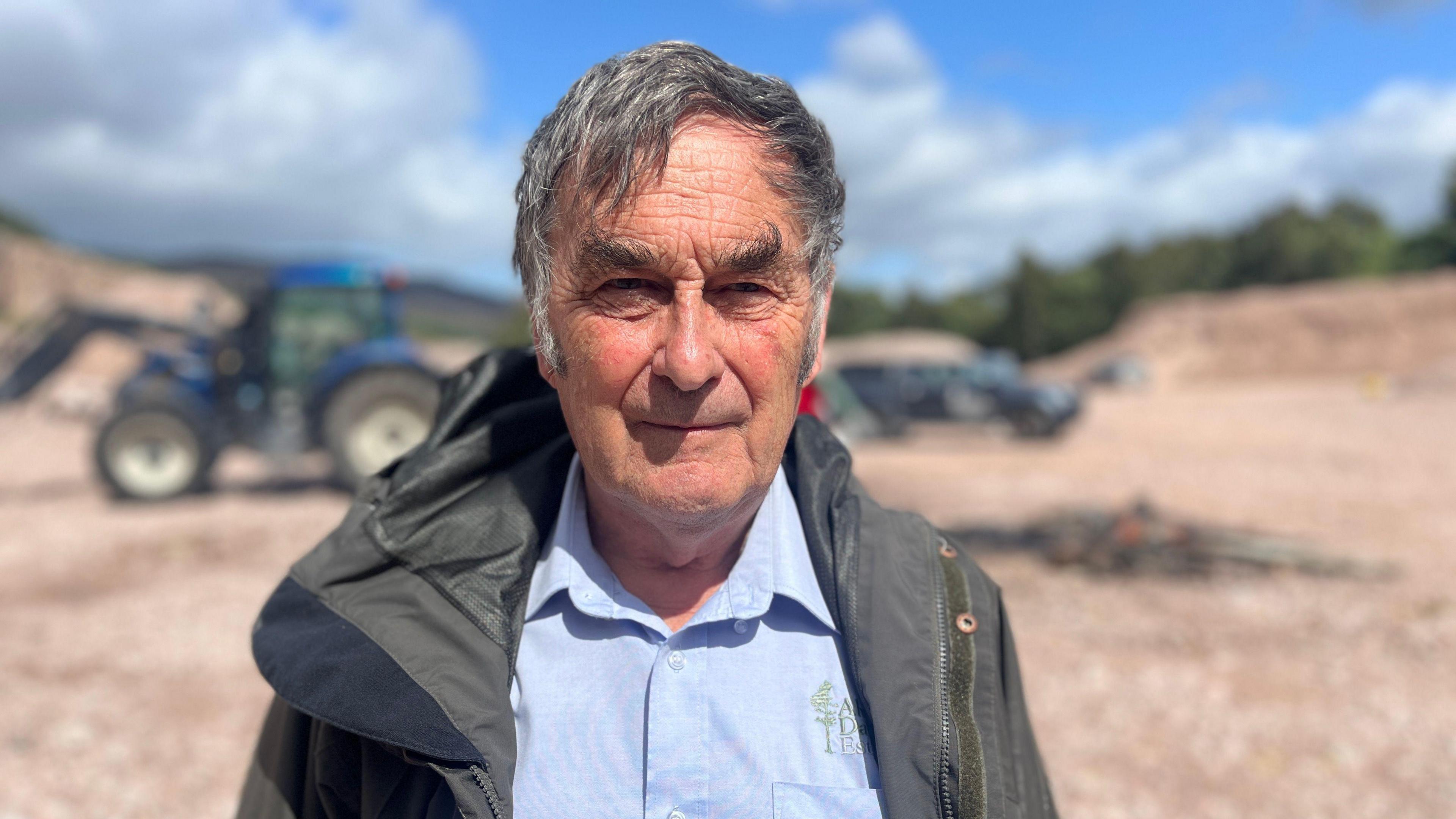 A man in a blue shirt and green jacket looks at the camera in front of a blue tractor and the remnants of a small fire.