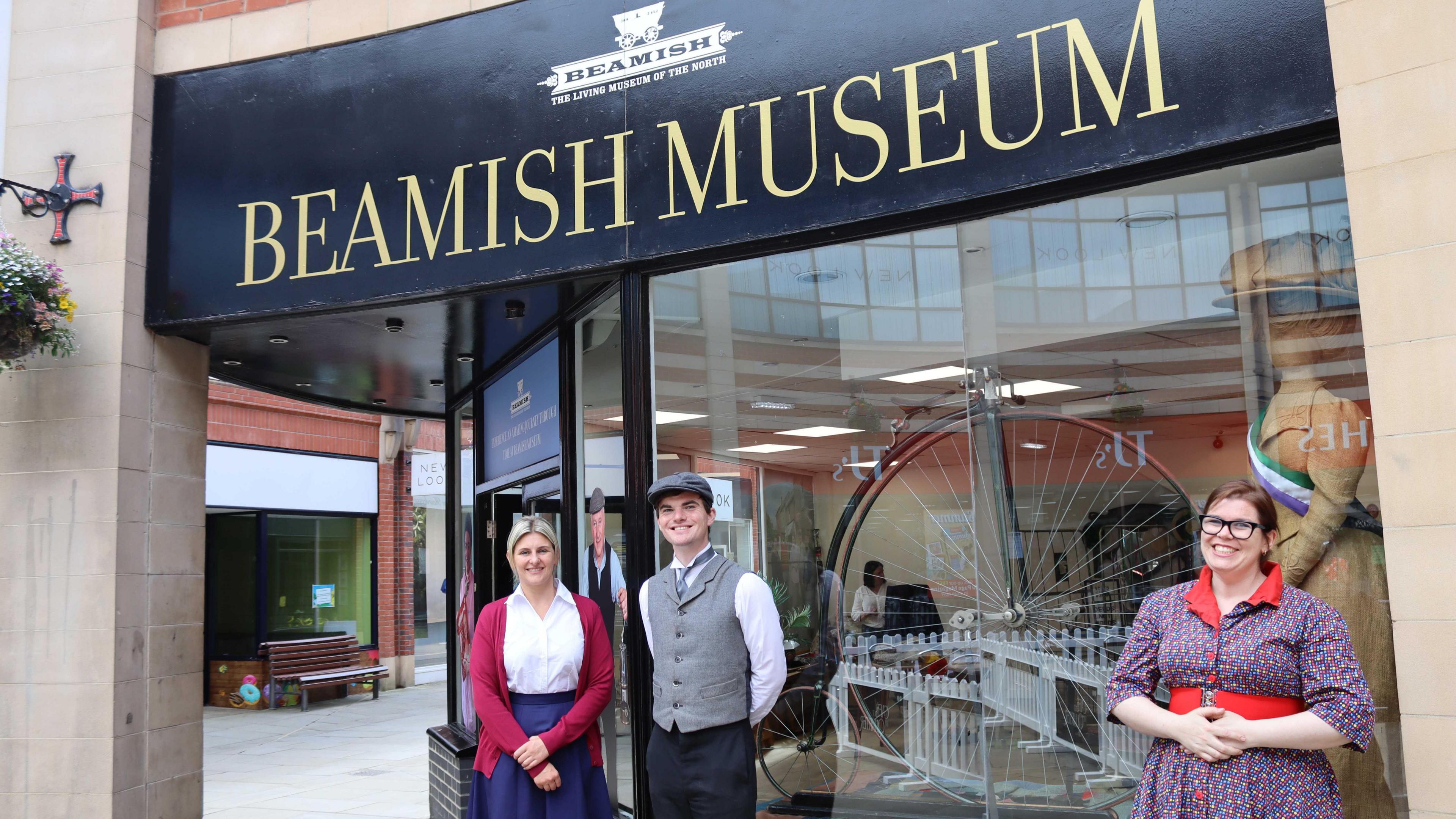 Rhiannon Hiles, Beamish Museum's CEO, stands with two members of staff wearing old-fashioned clothing outside of the shop. A penny-farthing can be seen in the window.