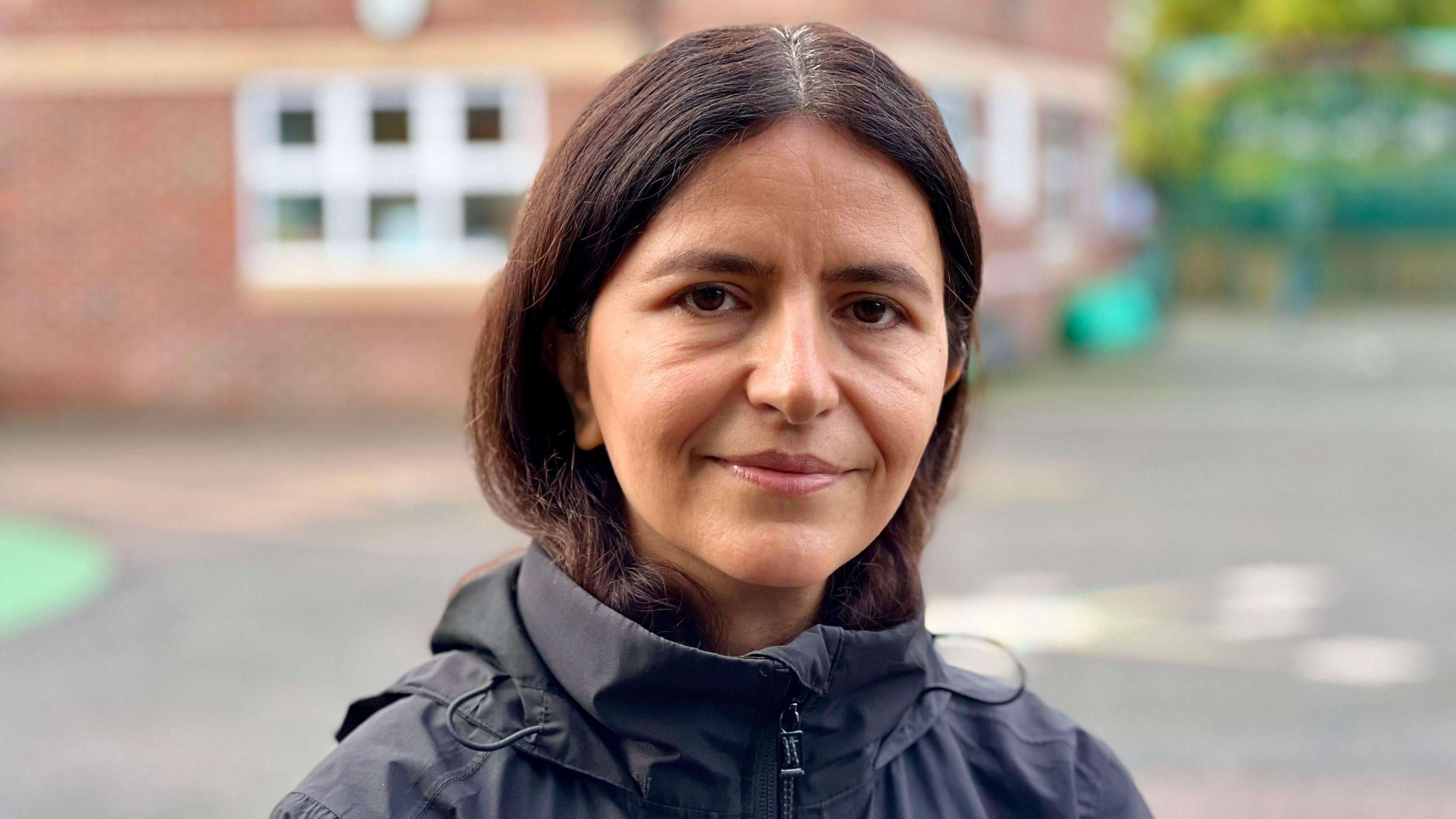 A picture of Shabnam looking and smiling at the camera on the school playground. Her dark her is tucked into a black raincoat. 