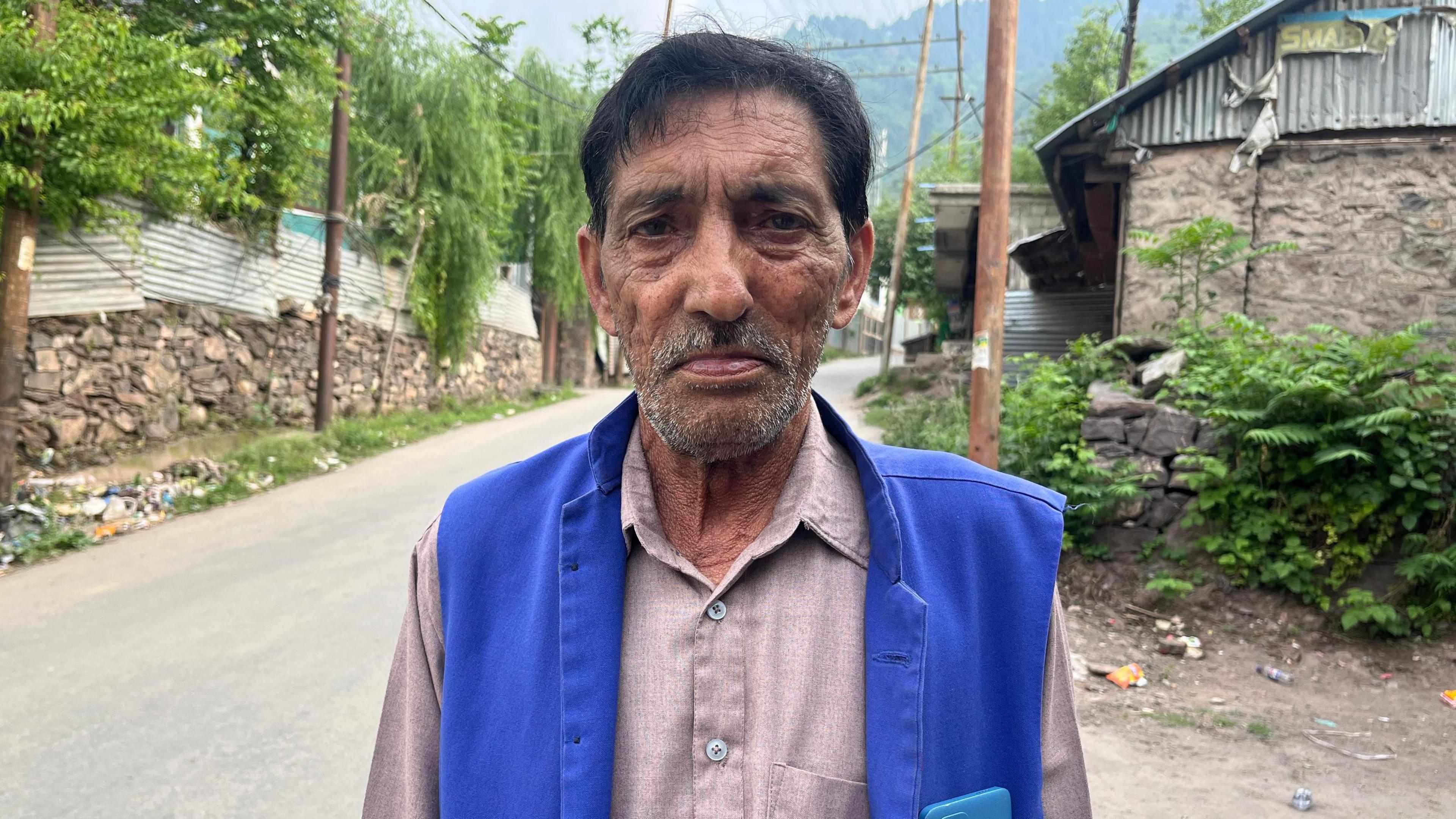 An elderly man stands on a road and looks into the camera, wearing traditional garb, homes made of stone and tin behind him