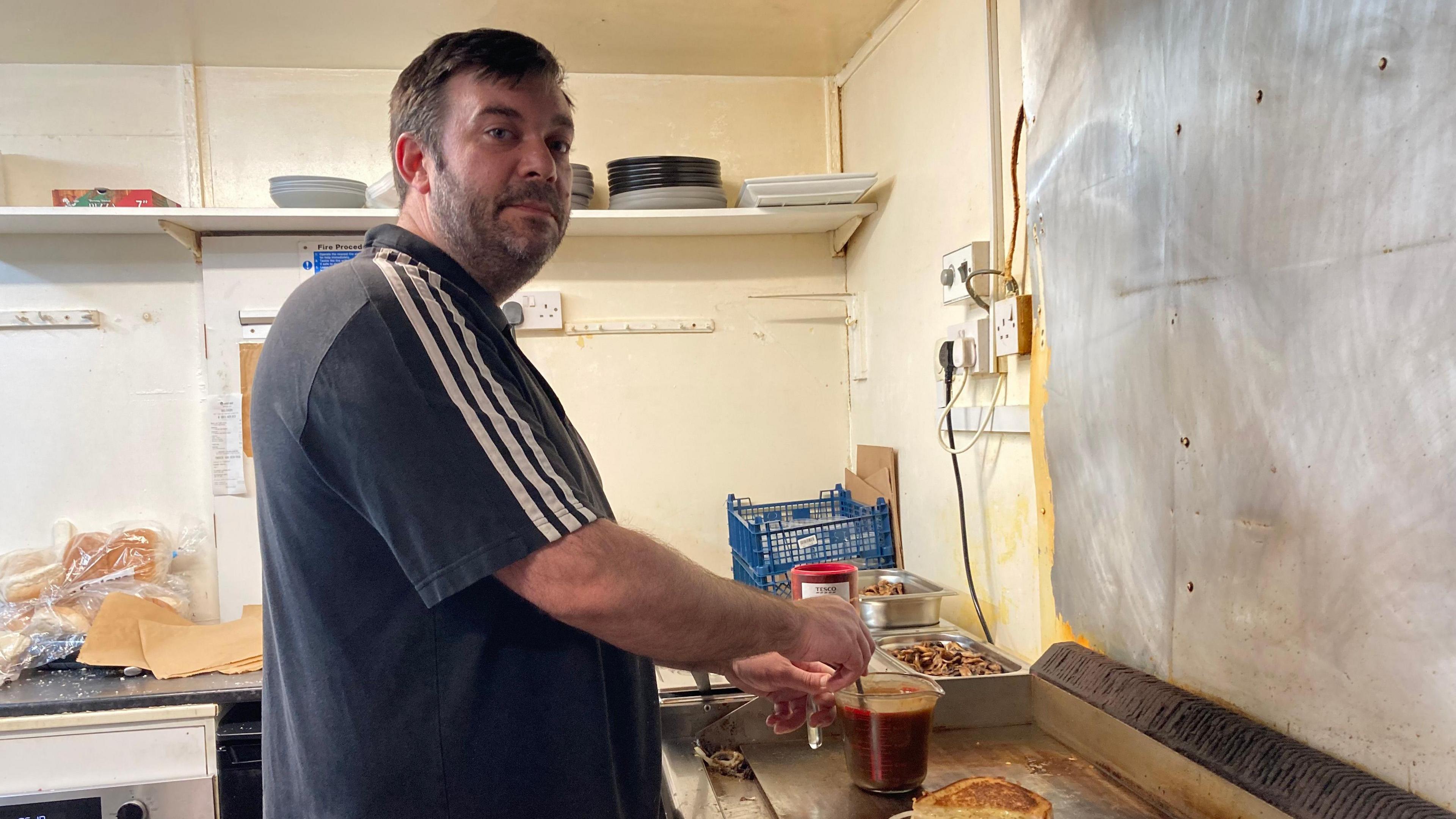 Craig Beverley, the owner of Bankside Cafe, is stood in a kitchen and stirs a jug filled with hot liquid as he looks at the camera. Mr Beverley has dark brown hair, a cropped beard and moustache. He is wearing a navy blue T-shirt with three white track lines on the sleeve.