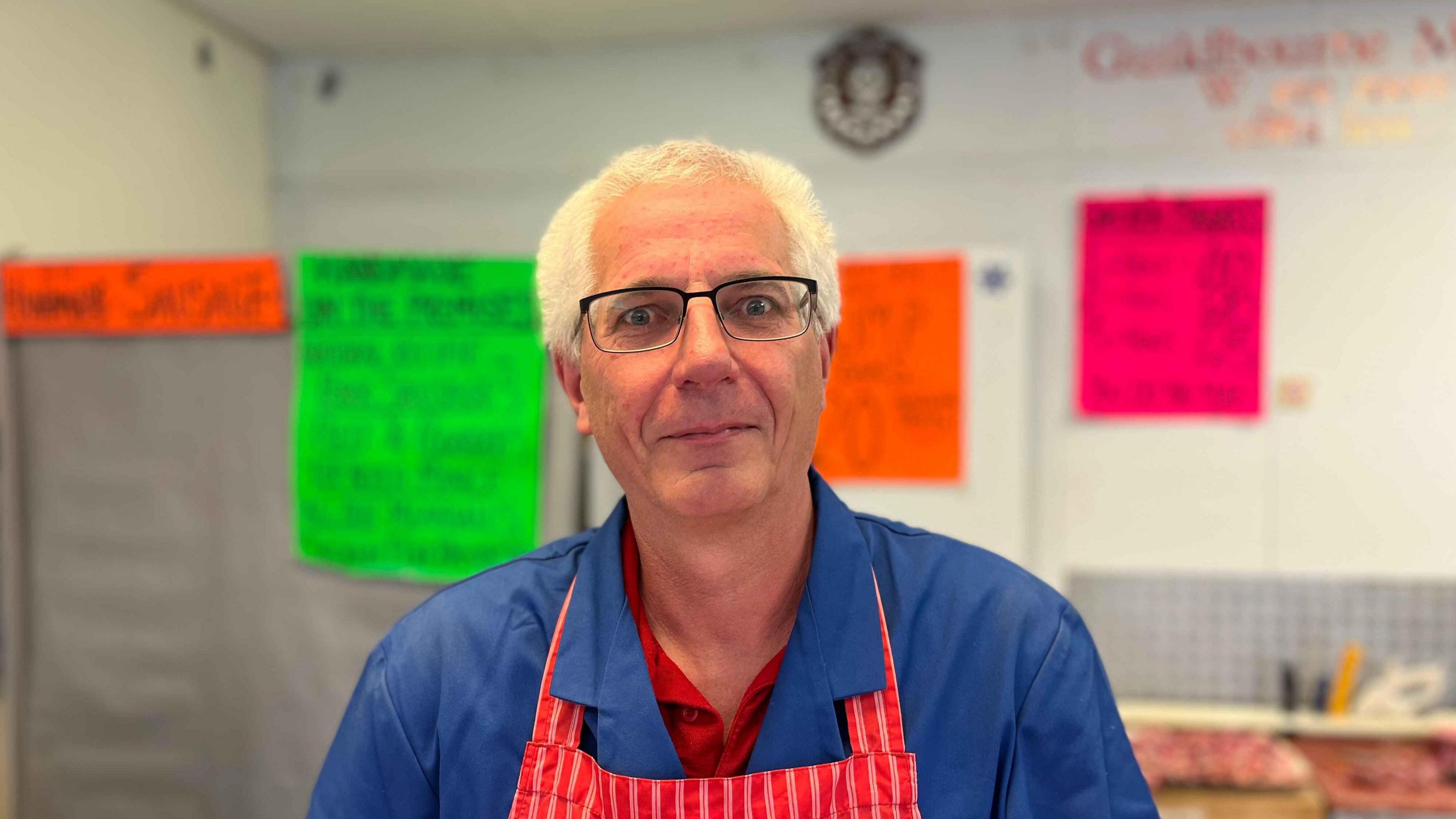 A head and shoulders shot of a man in a blue shirt, with a striped red apron. He has grey hair and is wearing black glasses. He is looking straight at the camera and there are fluorescent paper signs on the wall behind him