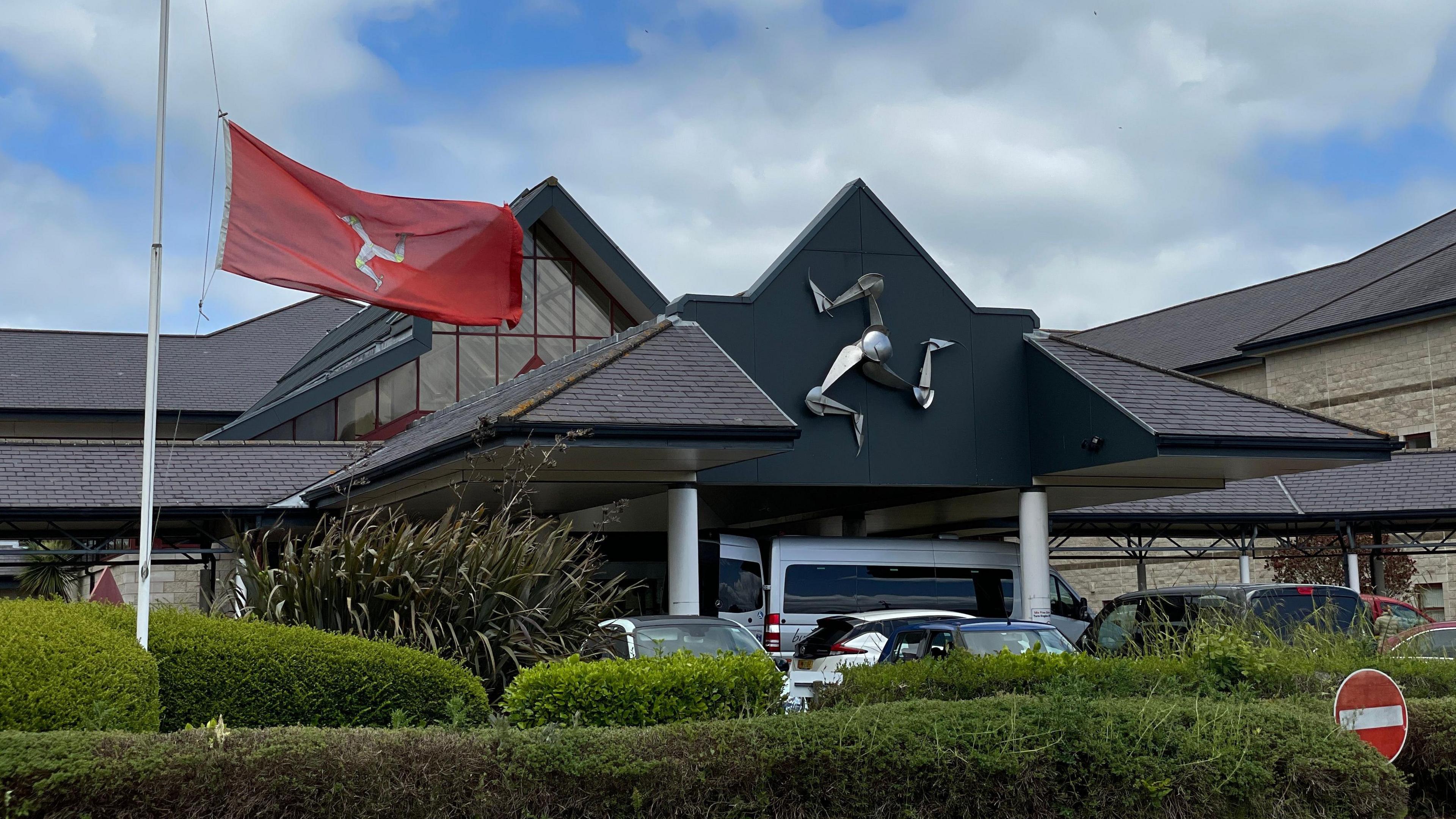 The exterior of the entrance to Noble's Hospital, which has a peaked roof with a metal triskelion sculpture on the front of it. There are cars are parked outside and a Manx flag on a white pole is blowing in the wind in front of it.